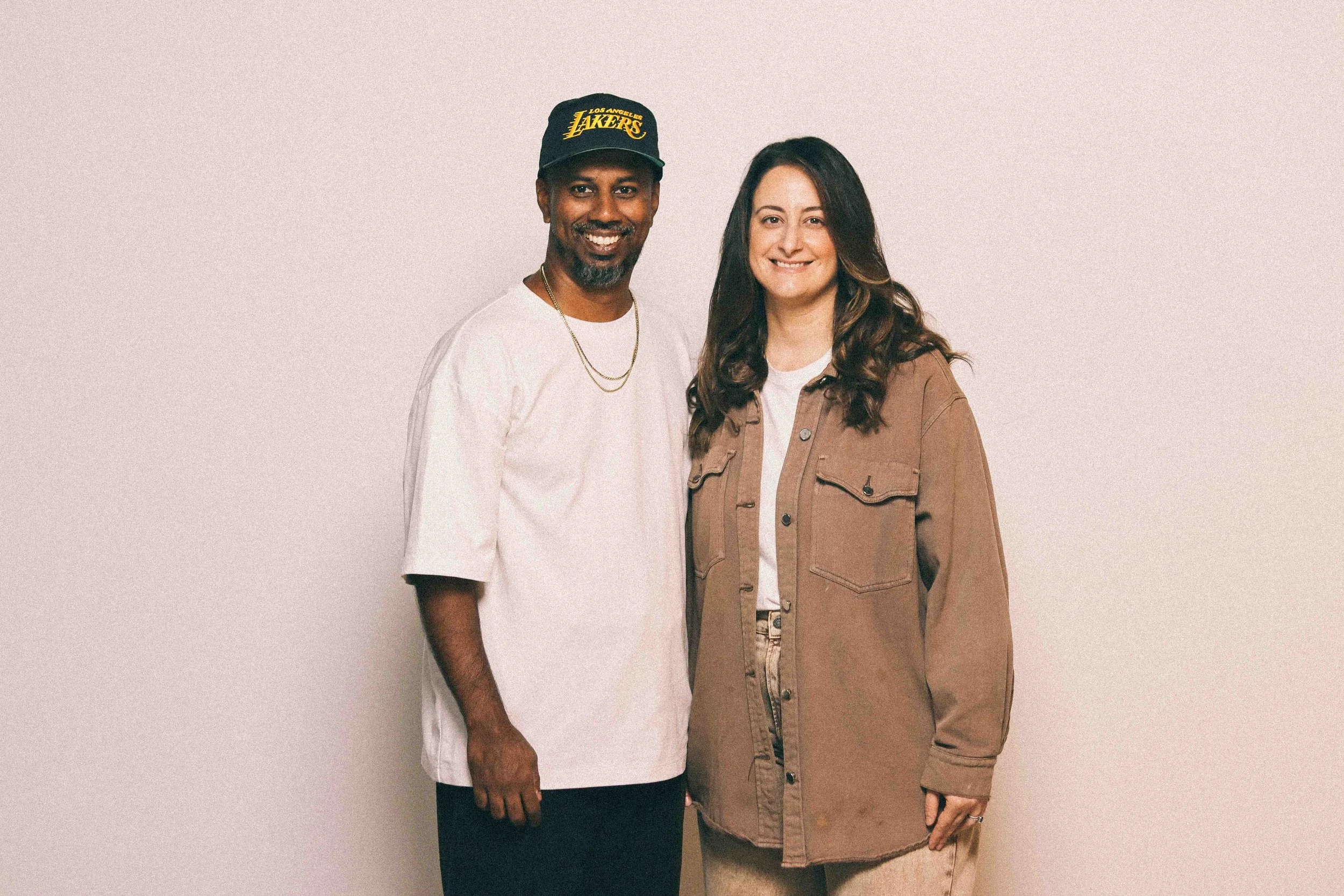 A man wearing a black Los Angeles Lakers baseball cap, white T-shirt, and gold chain standing next to a woman with long brown hair, wearing a brown jacket over a white shirt, both smiling and standing against a plain neutral background.
