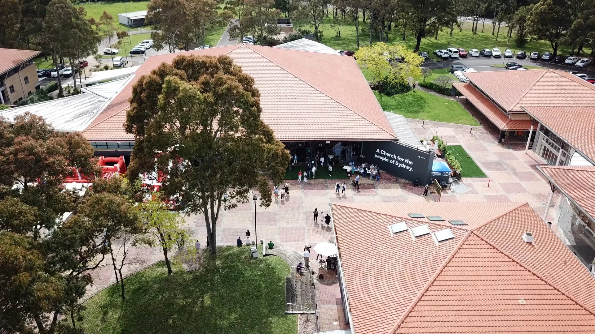 An aerial view of a church with groups of people entering and outside, surrounded by trees, buildings, and a parking lot.