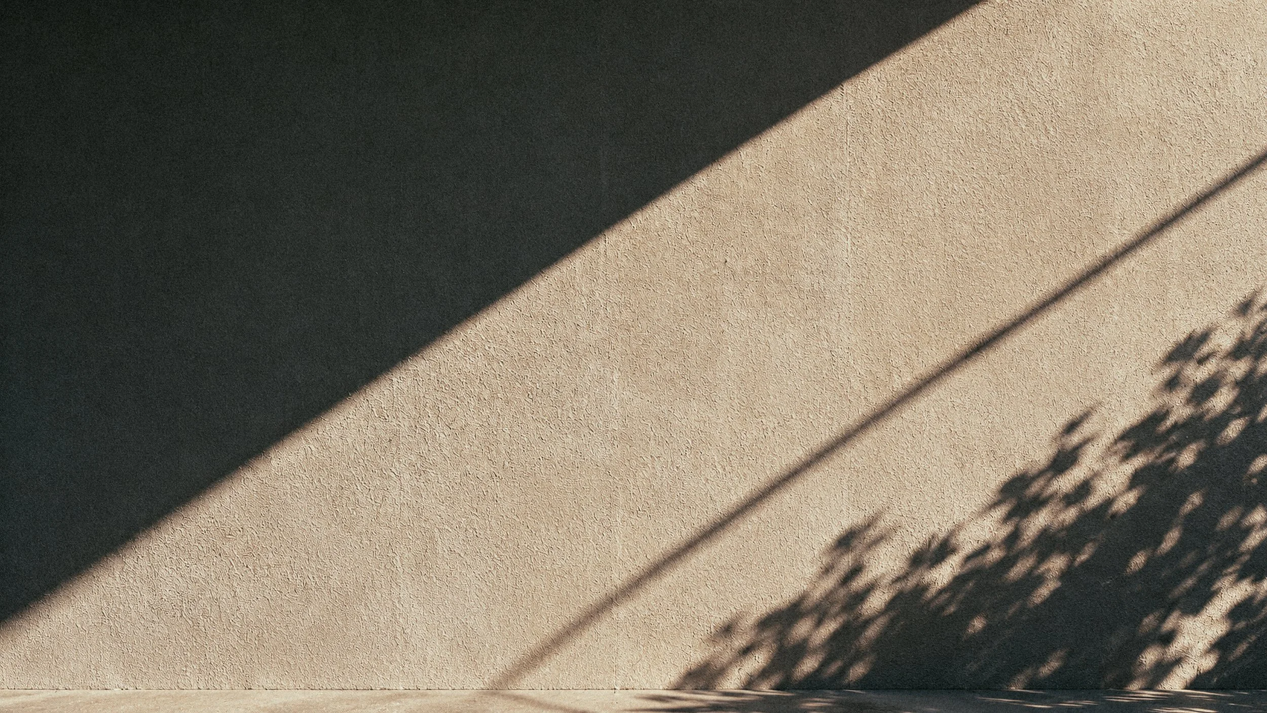 Shadow of a fence cast on a beige stucco wall, with the shadow creating a diagonal pattern.