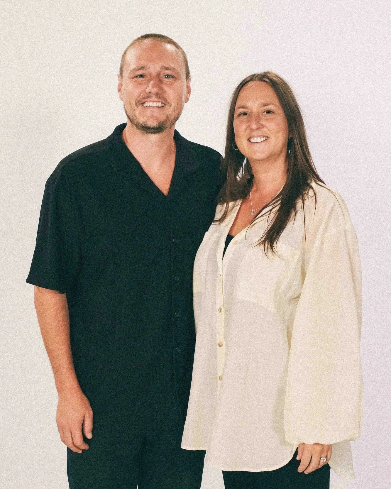 A man and woman standing together against a plain white wall, smiling at the camera.