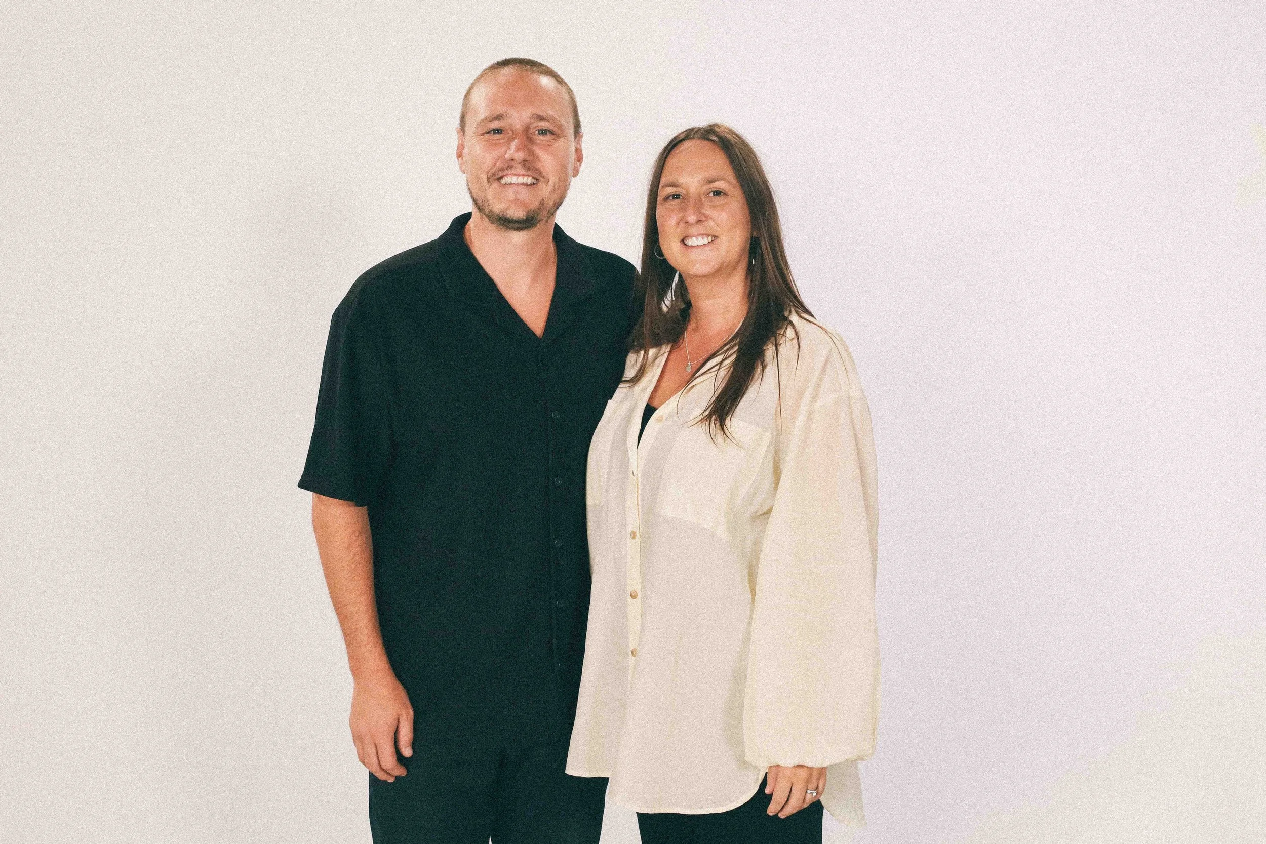 A man and woman standing close together, smiling, in front of a plain white background. The man has short hair, a beard, and is wearing a black shirt. The woman has long brown hair, is wearing earrings, a necklace, and a light-colored top.
