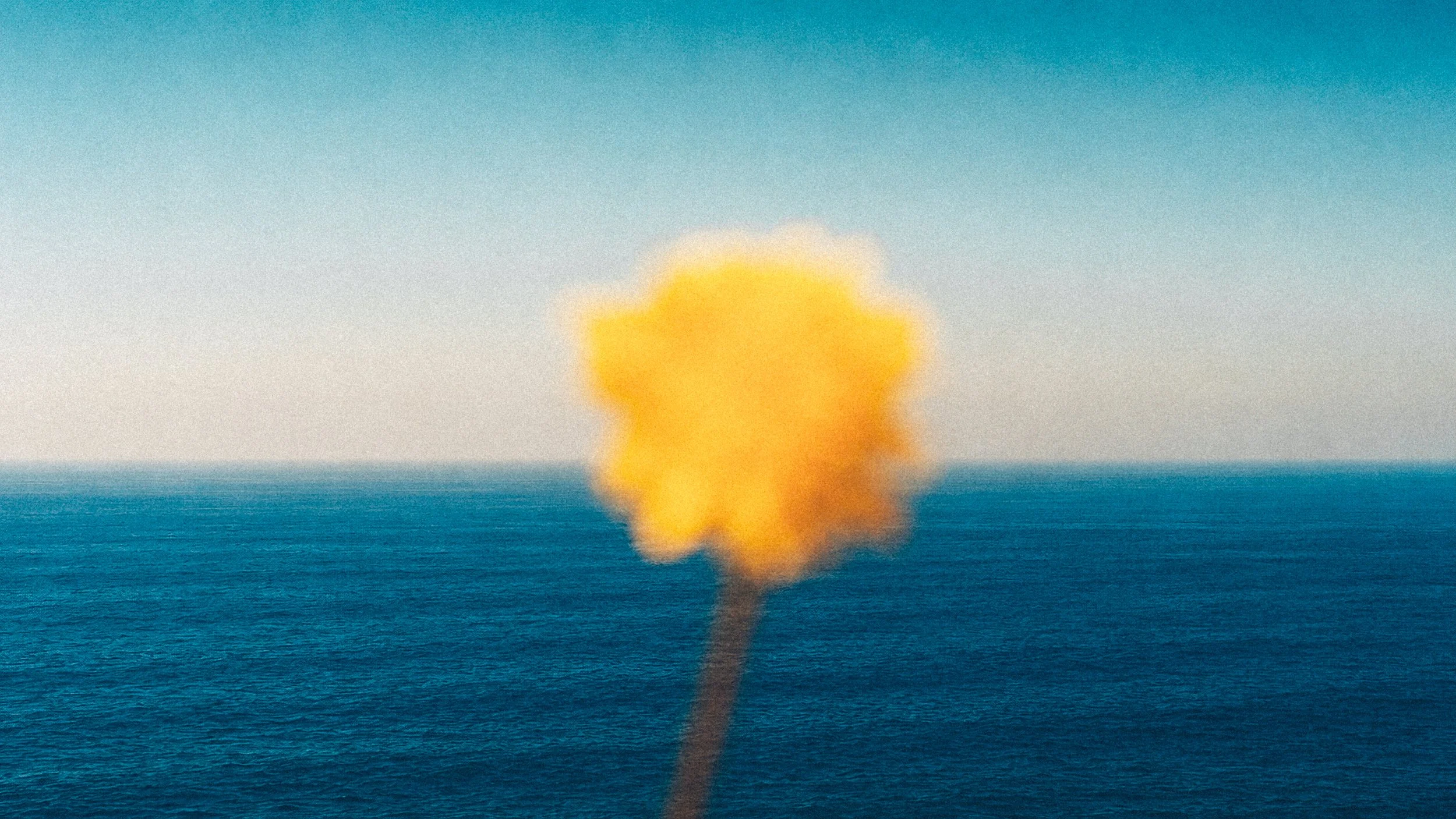 An explosion or large fire on the ocean with a bright yellow and orange plume of smoke rising into the sky, viewed from a distance.