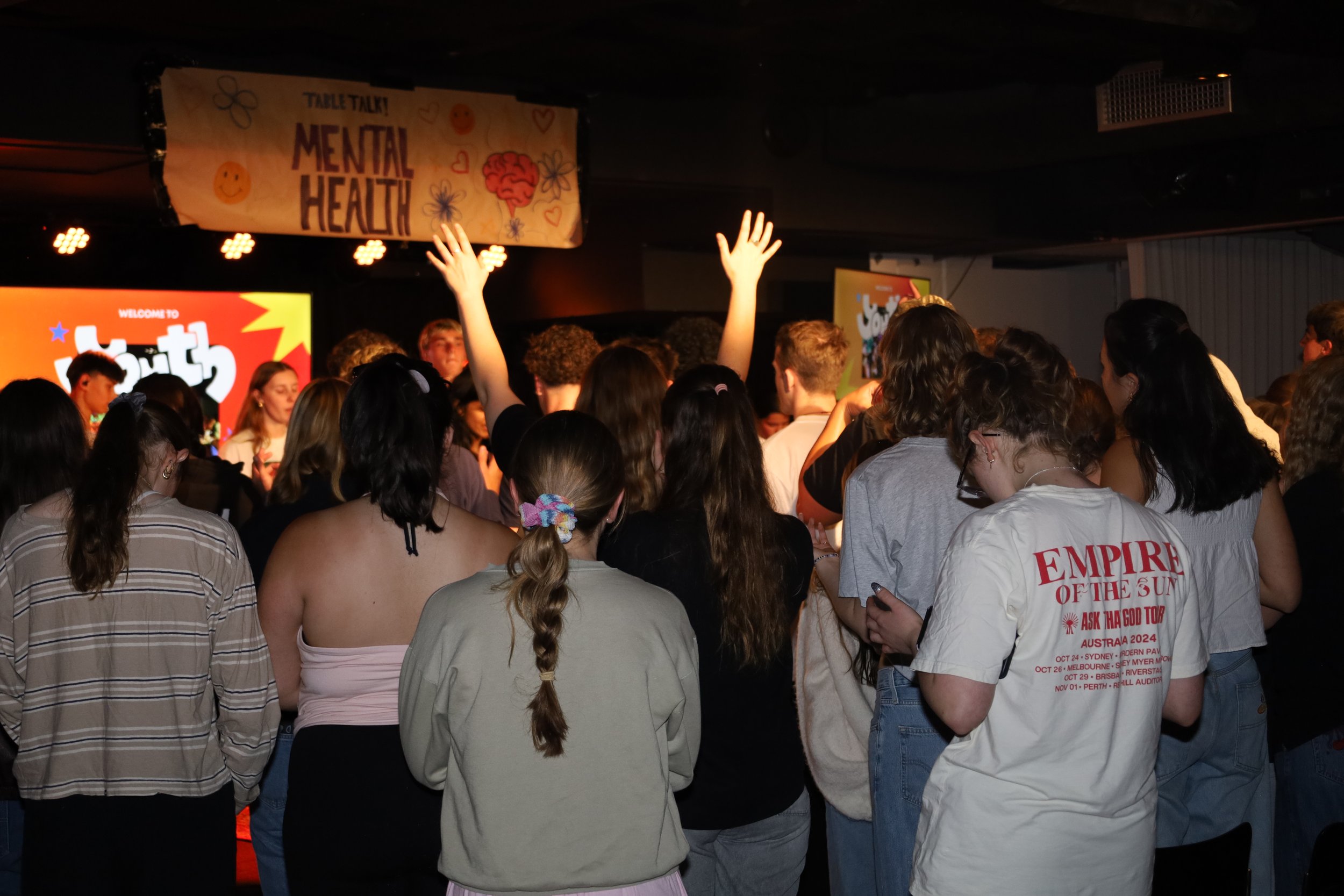 A large group of young people gathered at an event, with some raising their hands and others looking at a stage. There are screens and a banner related to mental health in the background.
