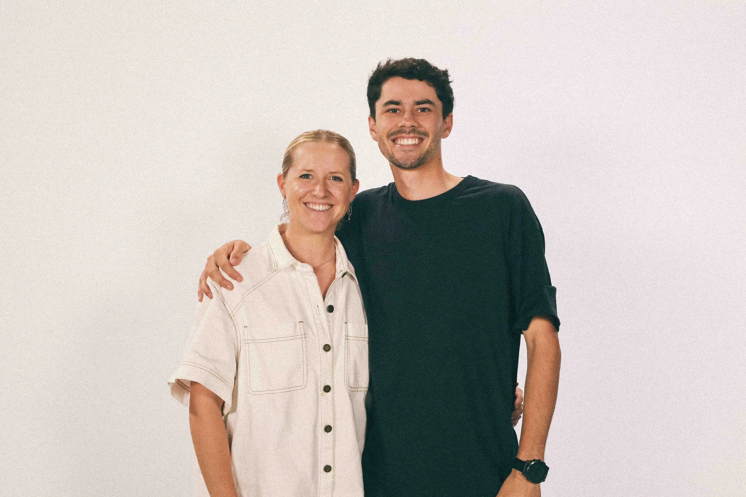 Smiling woman and man standing close together against a plain white background, with the woman on the left wearing a light-colored shirt and the man on the right wearing a black shirt.