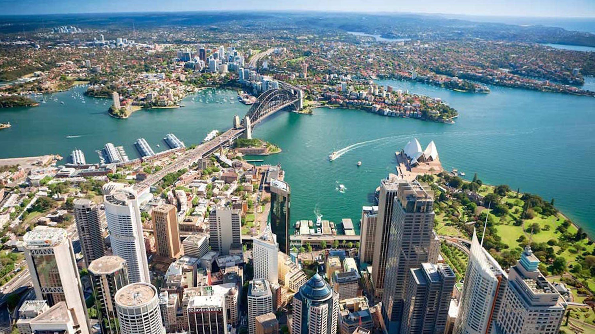 Aerial view of Sydney, Australia with the Sydney Opera House, Harbour Bridge, and Sydney Harbour, surrounded by high-rise buildings, green parks, and water with boats.