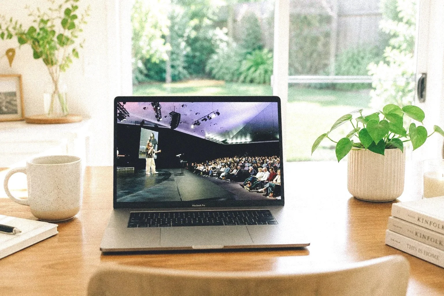 Laptop on a wooden desk showing a presentation to an audience, with a green potted plant to the right, a white mug, some books, and a notebook with a pen, near a large window with a garden view.