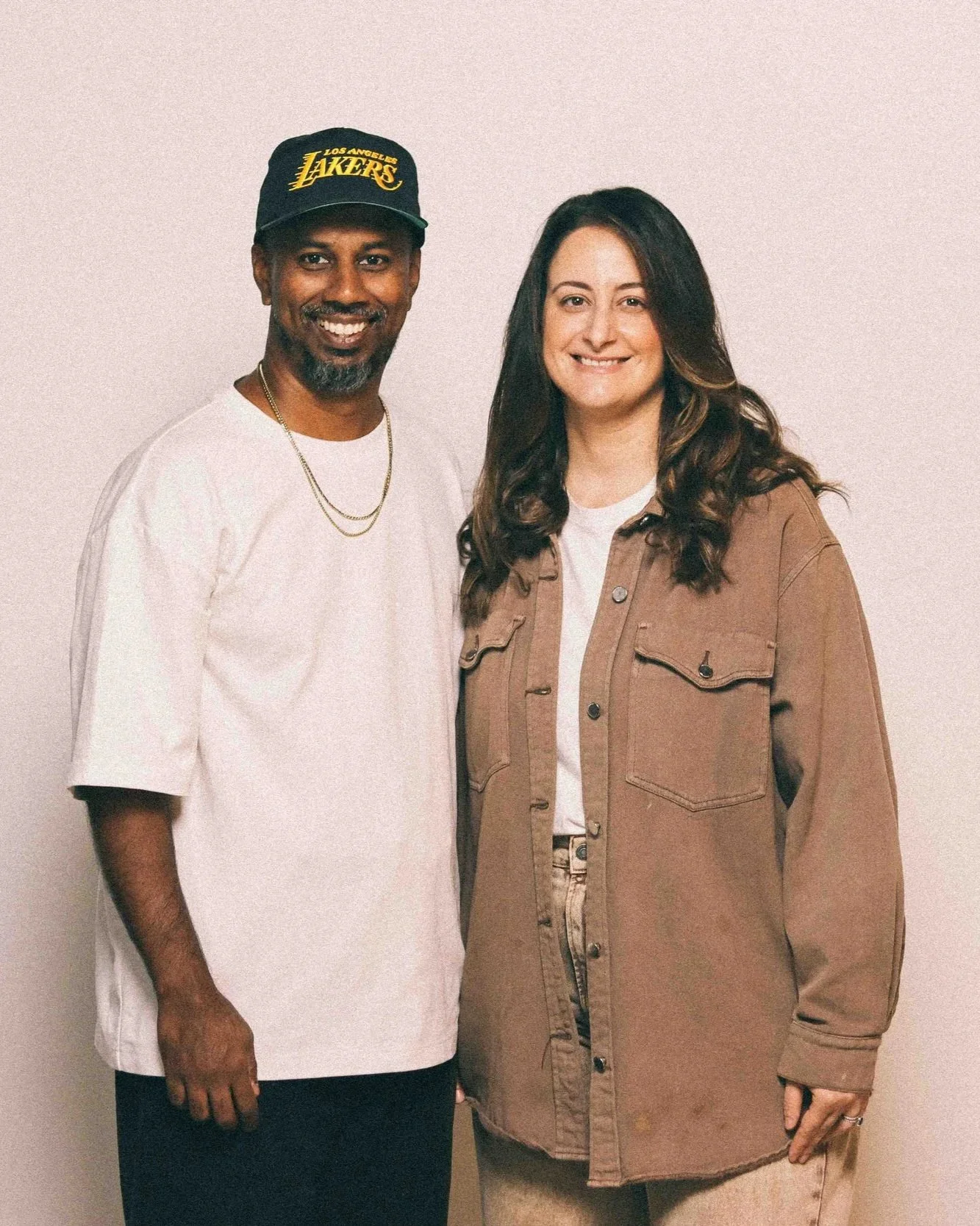 A man wearing a Los Angeles Lakers cap, white oversized t-shirt, and gold chains standing next to a woman with wavy dark hair, wearing a brown jacket and beige pants, both smiling at the camera against a plain wall.