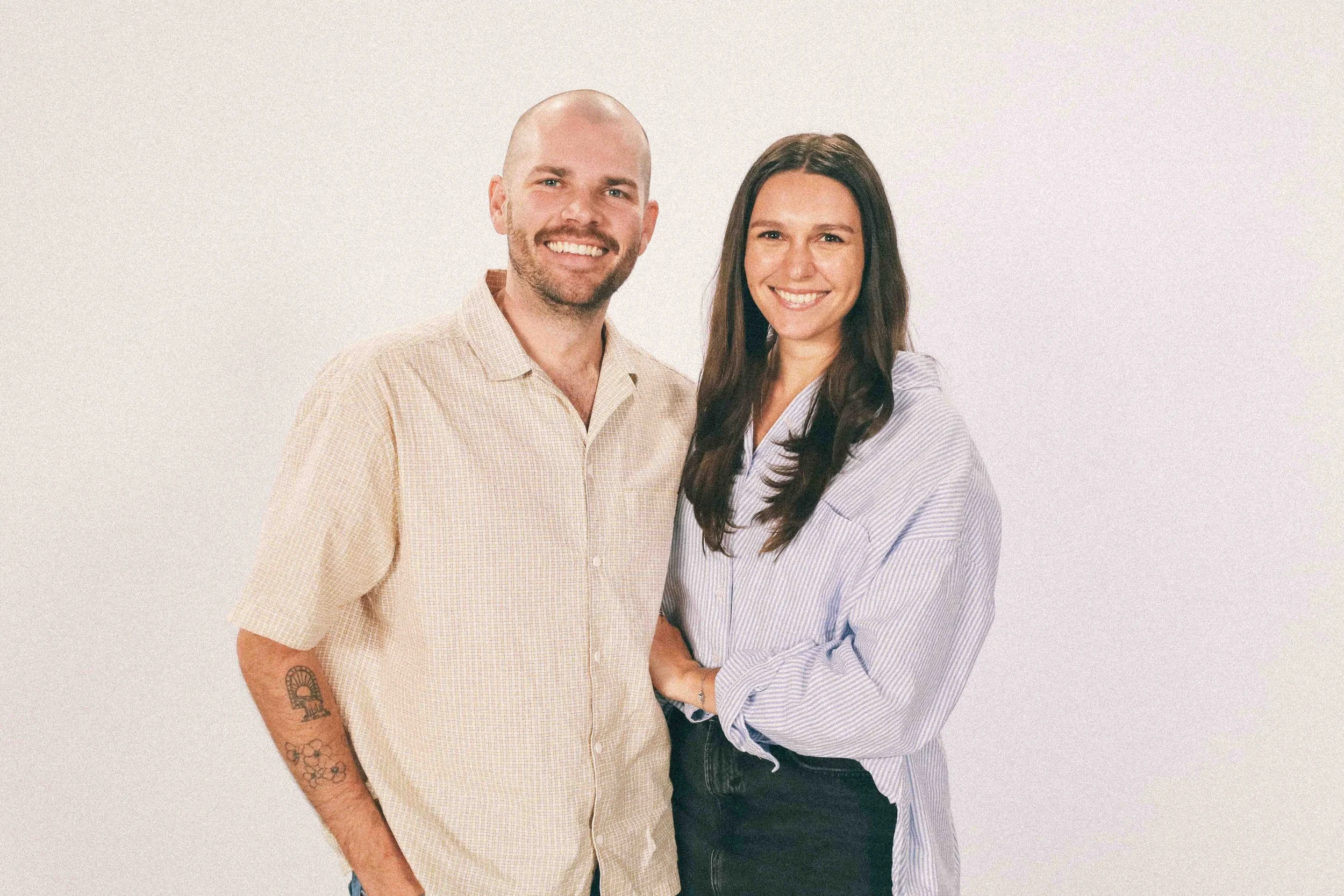 A man and woman smiling and standing close together against a white background, dressed casually in shirts.