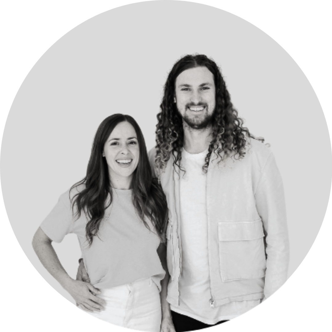 A black-and-white photo of a smiling woman and man standing close together against a plain background. The woman has long dark hair, and the man has long curly hair and a beard.