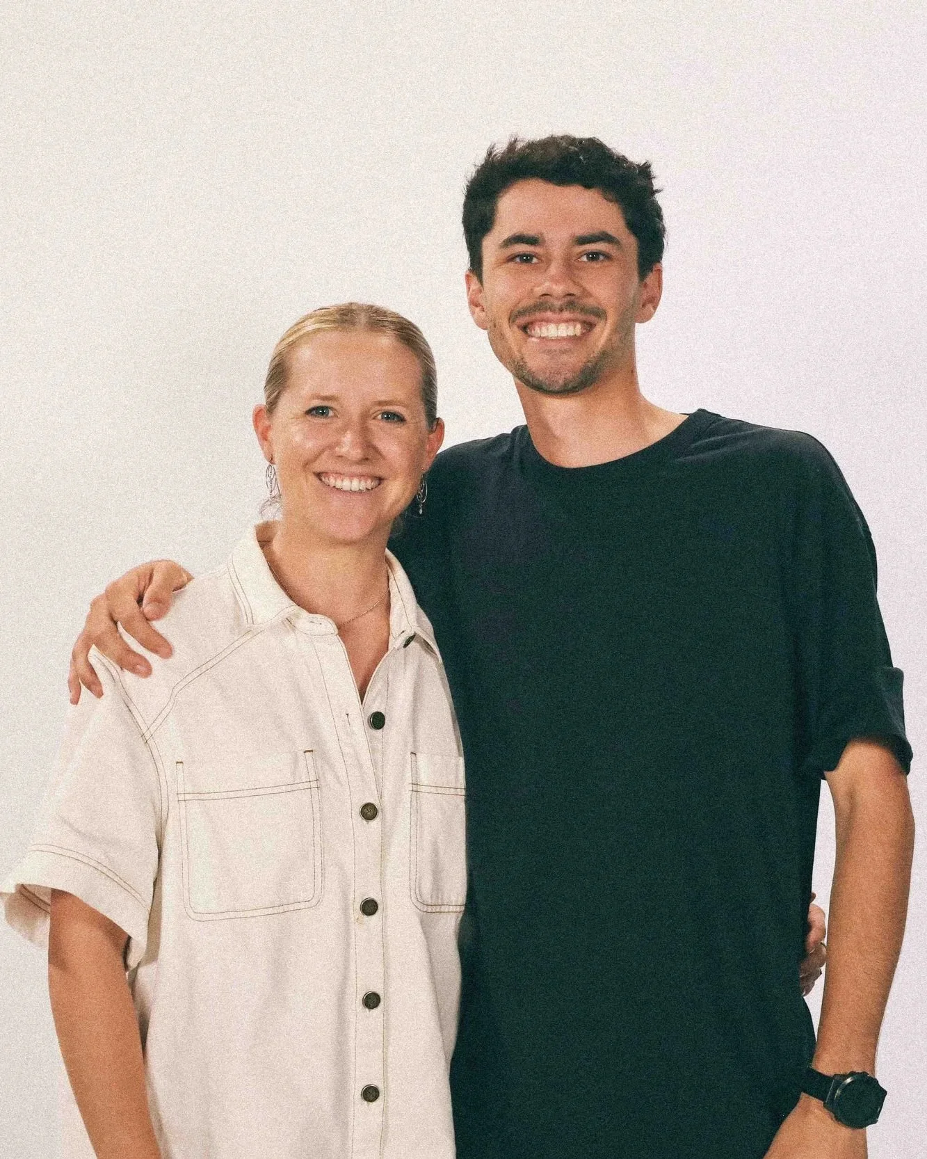 A woman with blonde hair and a man with dark hair standing together, smiling, with the woman's arm around the man's shoulder, against a white background.