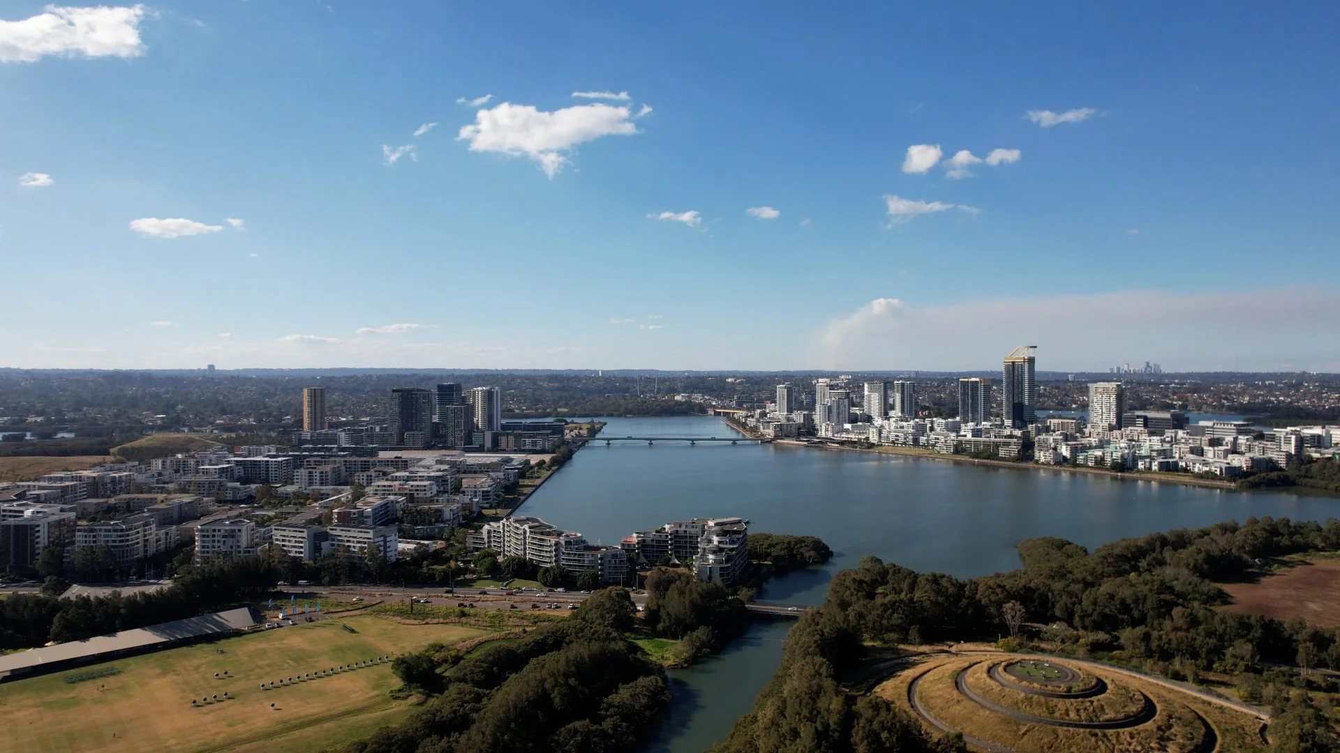 Aerial view of a city skyline with a river, high-rise buildings, and a park with walking paths and a roundabout, under a blue sky with fluffy clouds.