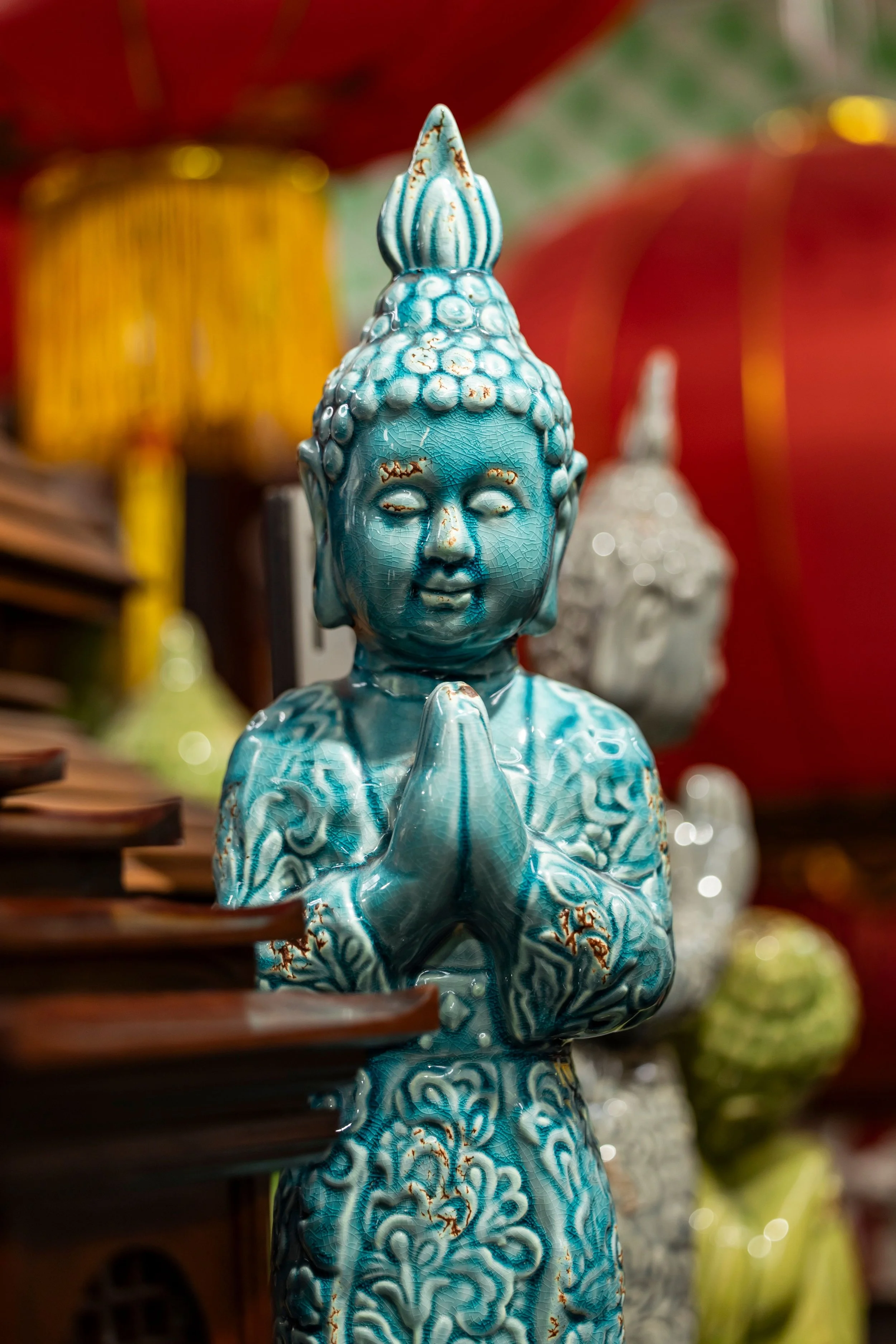 Statua in ceramica di Buddha in posizione di preghiera di colore blu con dettagli marroncini, sfondo sfocato con altri oggetti decorativi colorati.
