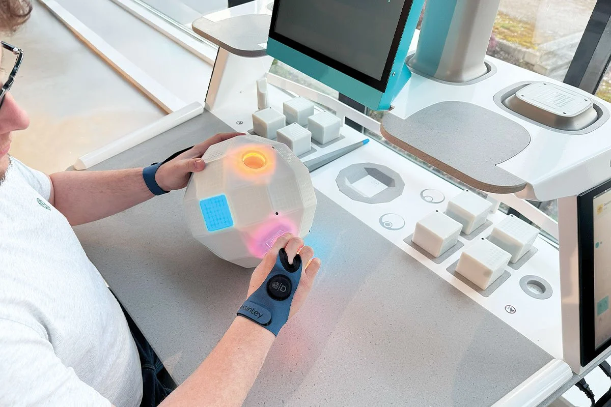 Person holding a white geodesic robot with colorful lights, sitting at a modern workstation with a computer, keypad, and lab equipment.
