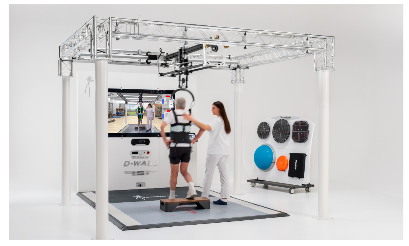 A person walking on a gait analysis treadmill with an exoskeleton device, assisted by a technician, in a clinical exercise testing lab with various equipment, including balance balls and training tools, in a bright white room.