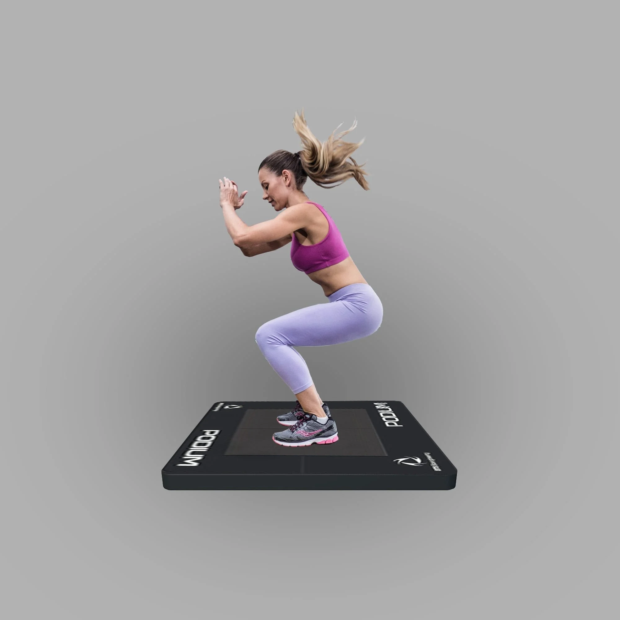 A woman wearing a pink sports bra and purple leggings jumping on a black exercise mat with white text, against a plain gray background.