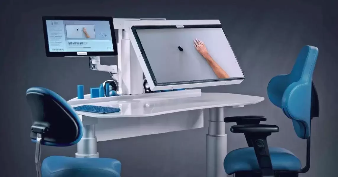Adaptive technology workspace with a large monitor displaying a hand writing on a virtual whiteboard, a computer keyboard, and multiple chairs including one with a specialized headrest.