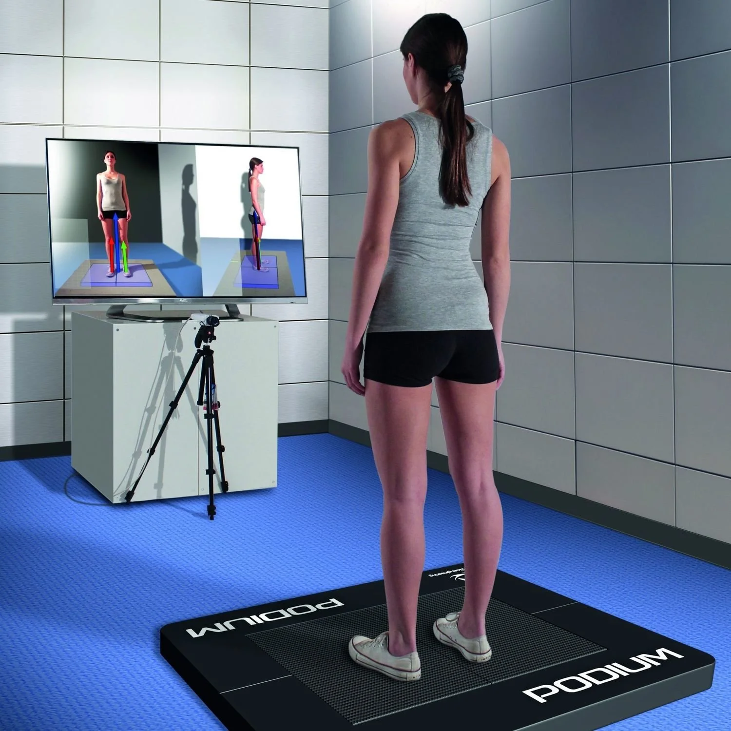 A woman stands on a Podium platform in a room with gray tiled walls, looking at a large screen displaying a 3D body model with arrows indicating movement. A camera on a tripod is positioned in front of her.
