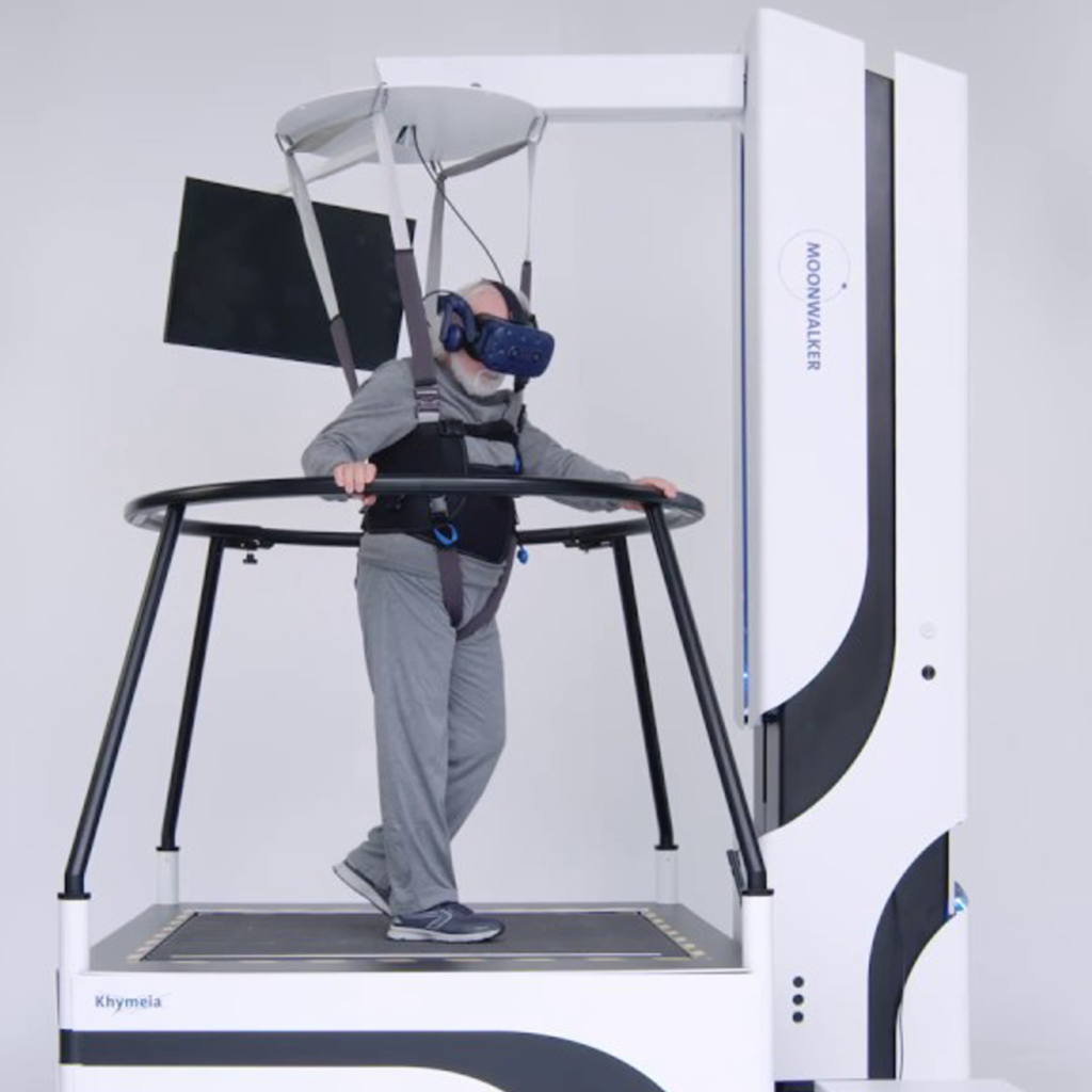 An elderly man wearing a VR headset and headphones, standing on a treadmill-like device within a simulated environment with a harness and support structure around him.