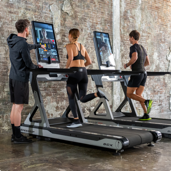 Three people are running on treadmills in a gym with brick walls. Two are looking at screens displaying a video call or workout info, and one person is running on a treadmill.