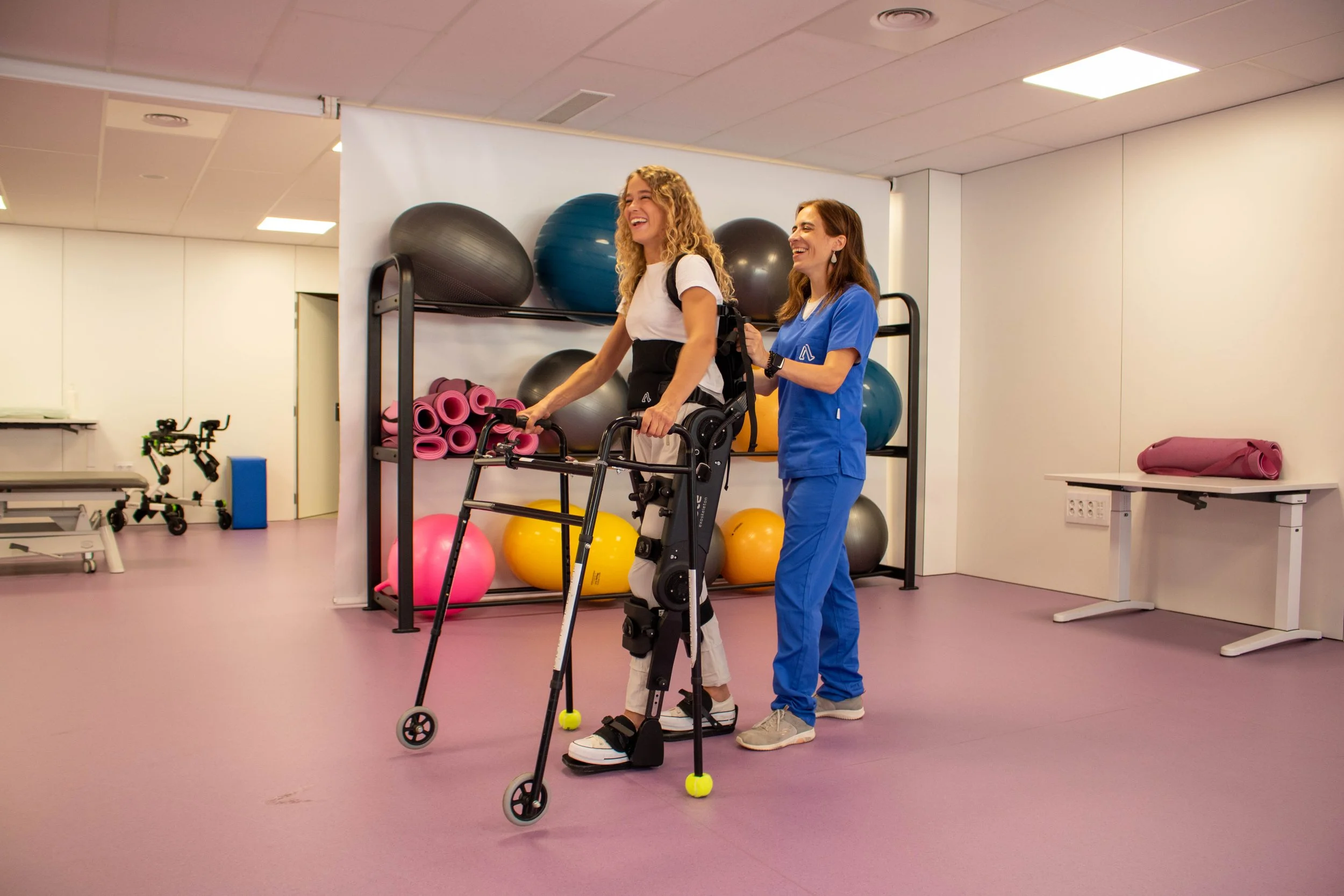 A young woman in a walker with a green tennis ball on the front foot is being assisted by a healthcare worker in blue scrubs inside a physiotherapy or rehabilitation center. They are both smiling and appear to be engaged in a therapy session. The background shows exercise balls, pink yoga mats, and other rehabilitation equipment.