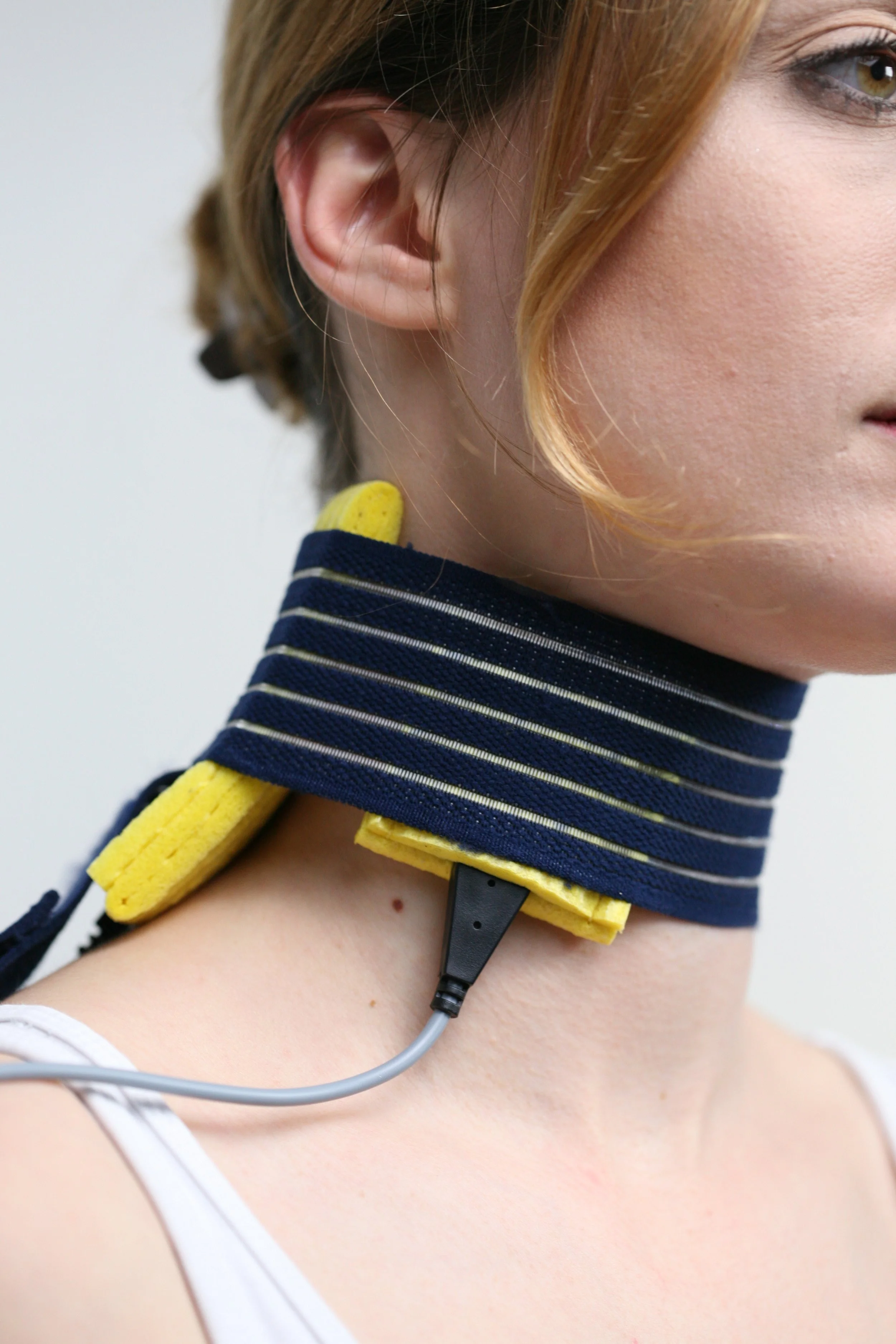 Close-up of a woman wearing a striped navy and white shirt, with a yellow padded necklace and a headset connected to a device. The woman's face is partially visible, showing her ear, eye, and cheek.