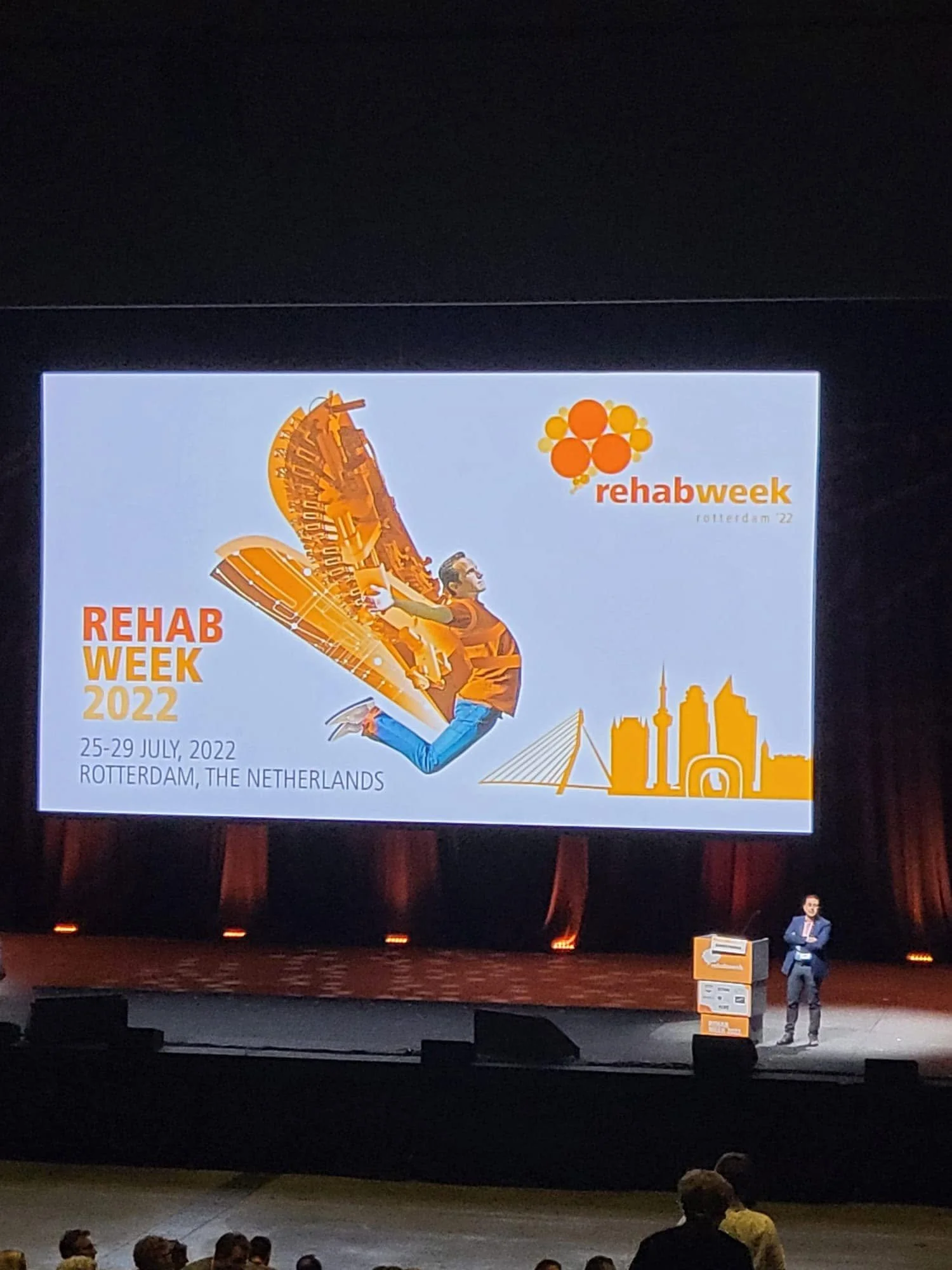 A large digital display advertising Rehab Week 2022 in Rotterdam, Netherlands, with a graphic of a person jumping in front of a rollercoaster and a city skyline.