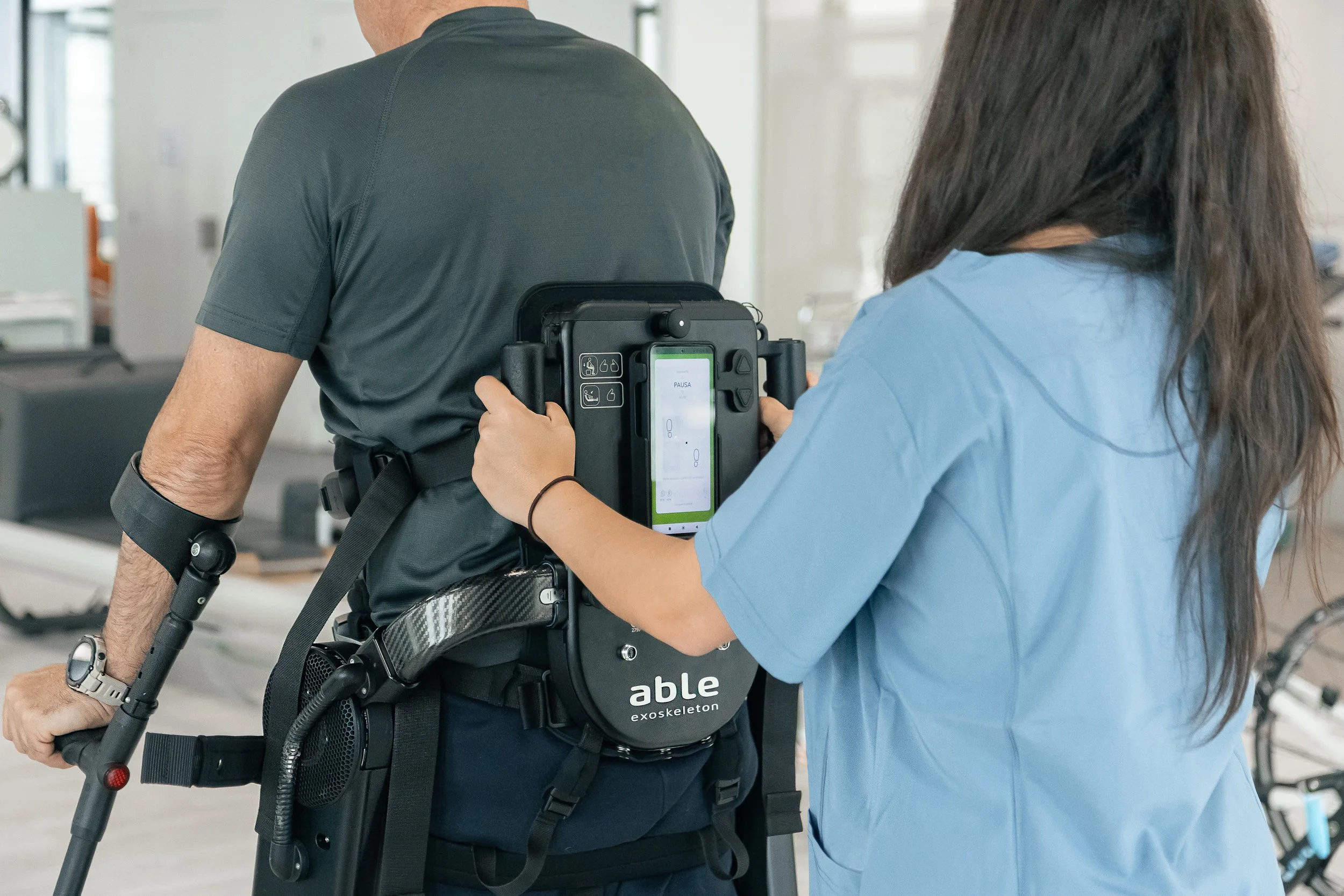 A healthcare worker in blue scrubs attaching a monitor to a patient who is walking with a gait trainer in a medical or rehabilitation facility.