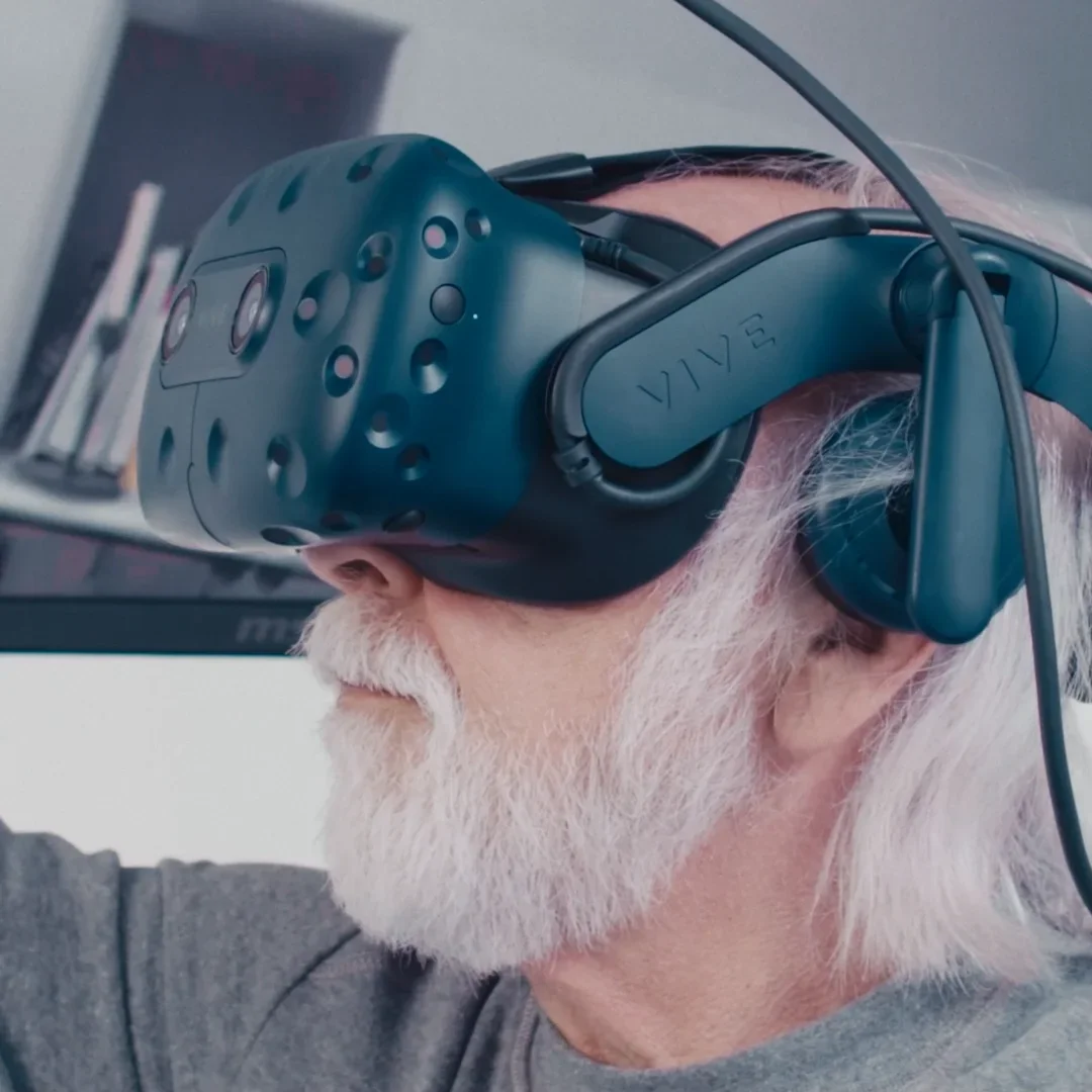 An elderly man with white hair and beard wearing a VR headset labeled VIVE.