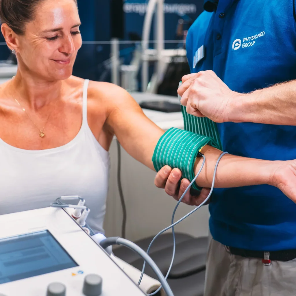 A woman receiving a medical test on her arm from a healthcare professional in a clinical setting. The professional is wearing a blue shirt with a logo, and the woman is smiling while looking at her arm.