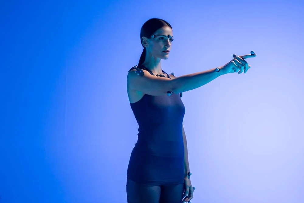 A woman dressed in a black sleeveless dress and cybernetic implants on her arms and shoulders, standing against a blue background, extending her right arm outward.