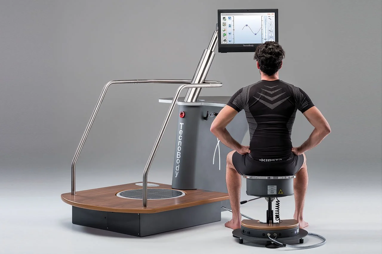 A man sitting on a rotating, padded seat connected to a medical or scientific device with a monitor displaying a graph, in a clinical setting.