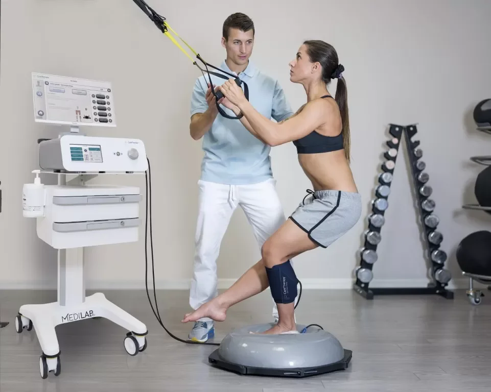 A woman in workout clothes standing on a balance platform, holding resistance bands while a male trainer assists her during a rehabilitation session in a gym setting with medical equipment and weights.