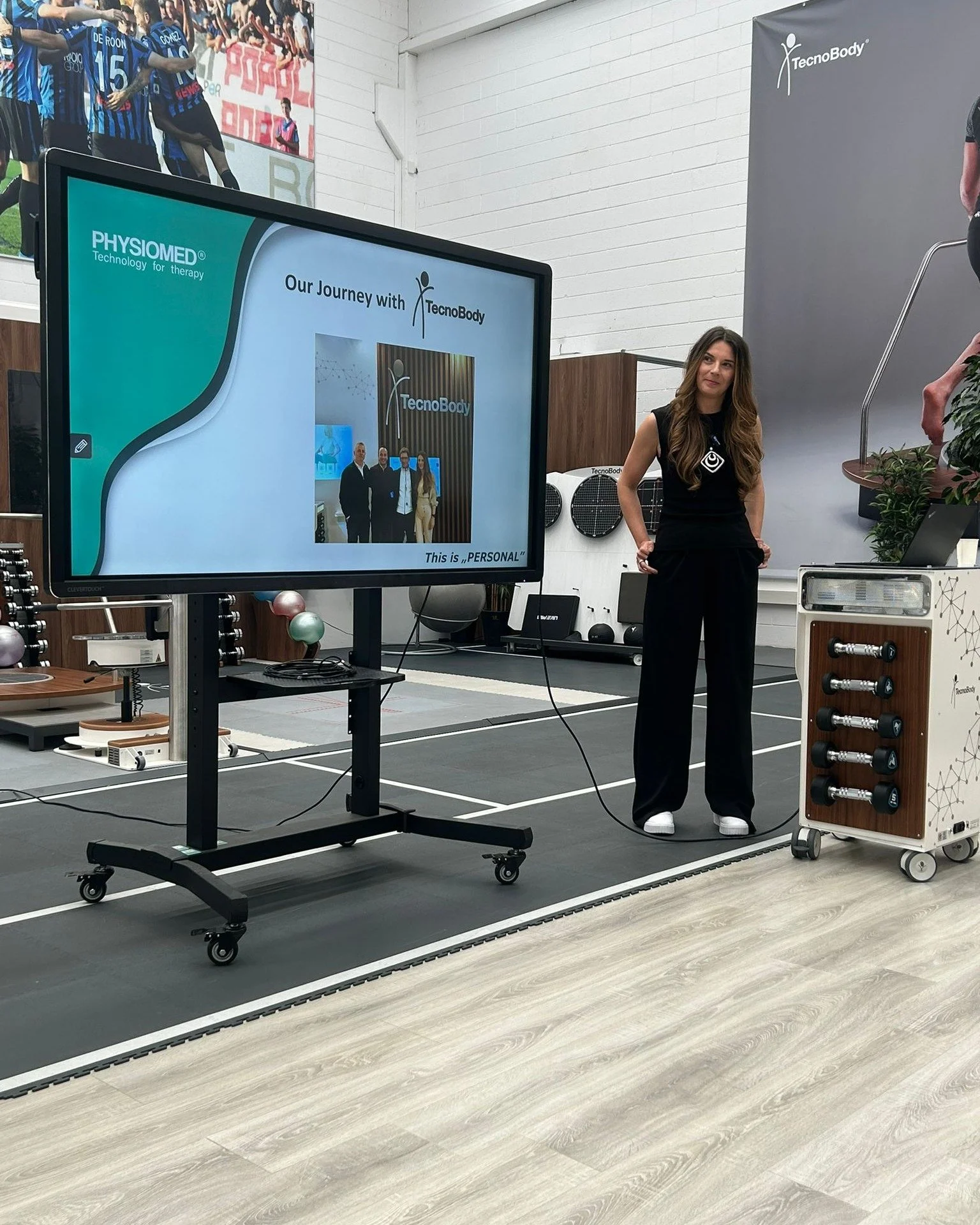 Woman standing with hands on hips next to a display screen at a health or fitness event, with a banner and posters related to TecnoBody and PhysioMed in the background.