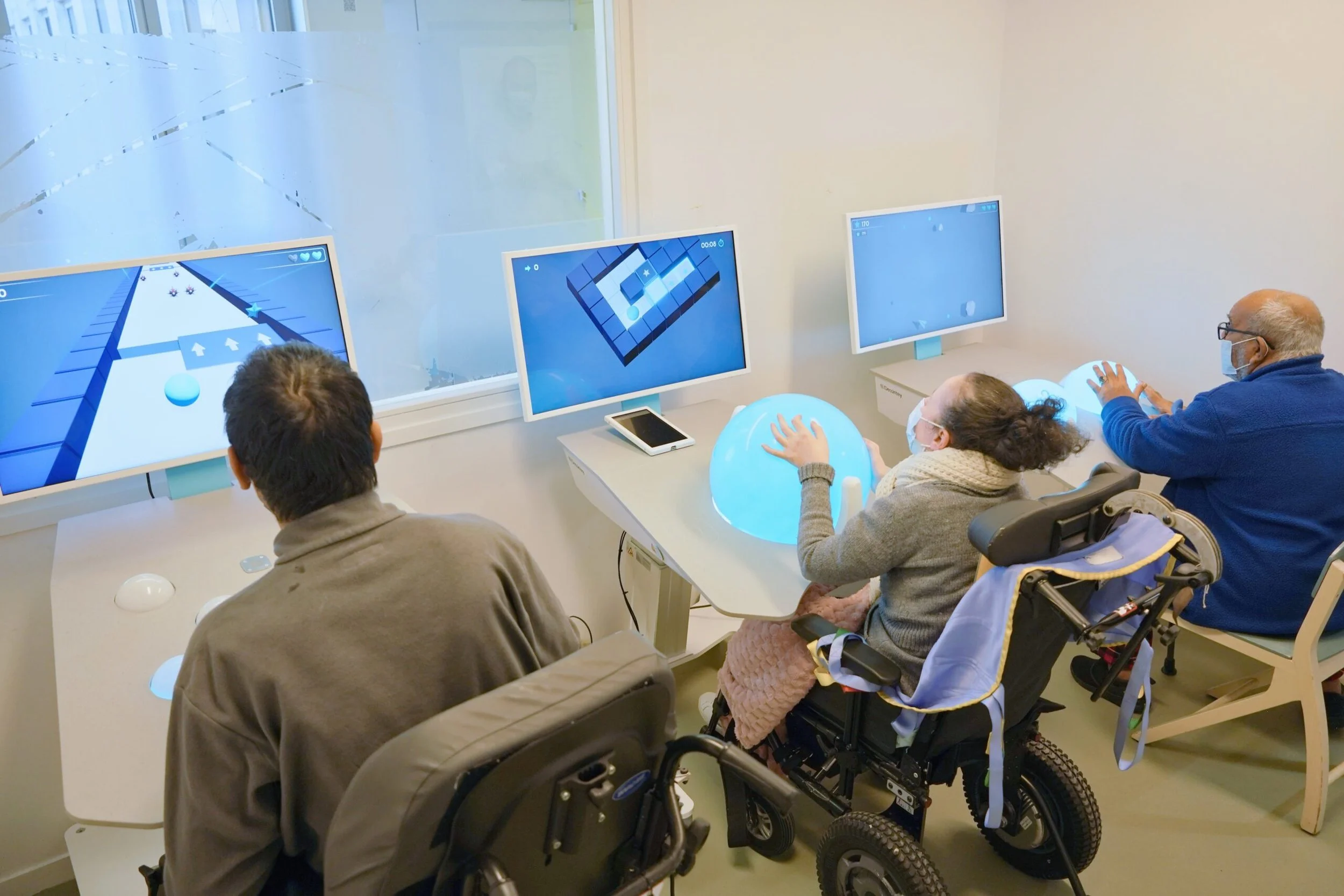 People playing a video game on computers with large monitors in a well-lit room. Some players are in wheelchairs. A woman is holding a blue exercise ball, and others are focused on the screens.