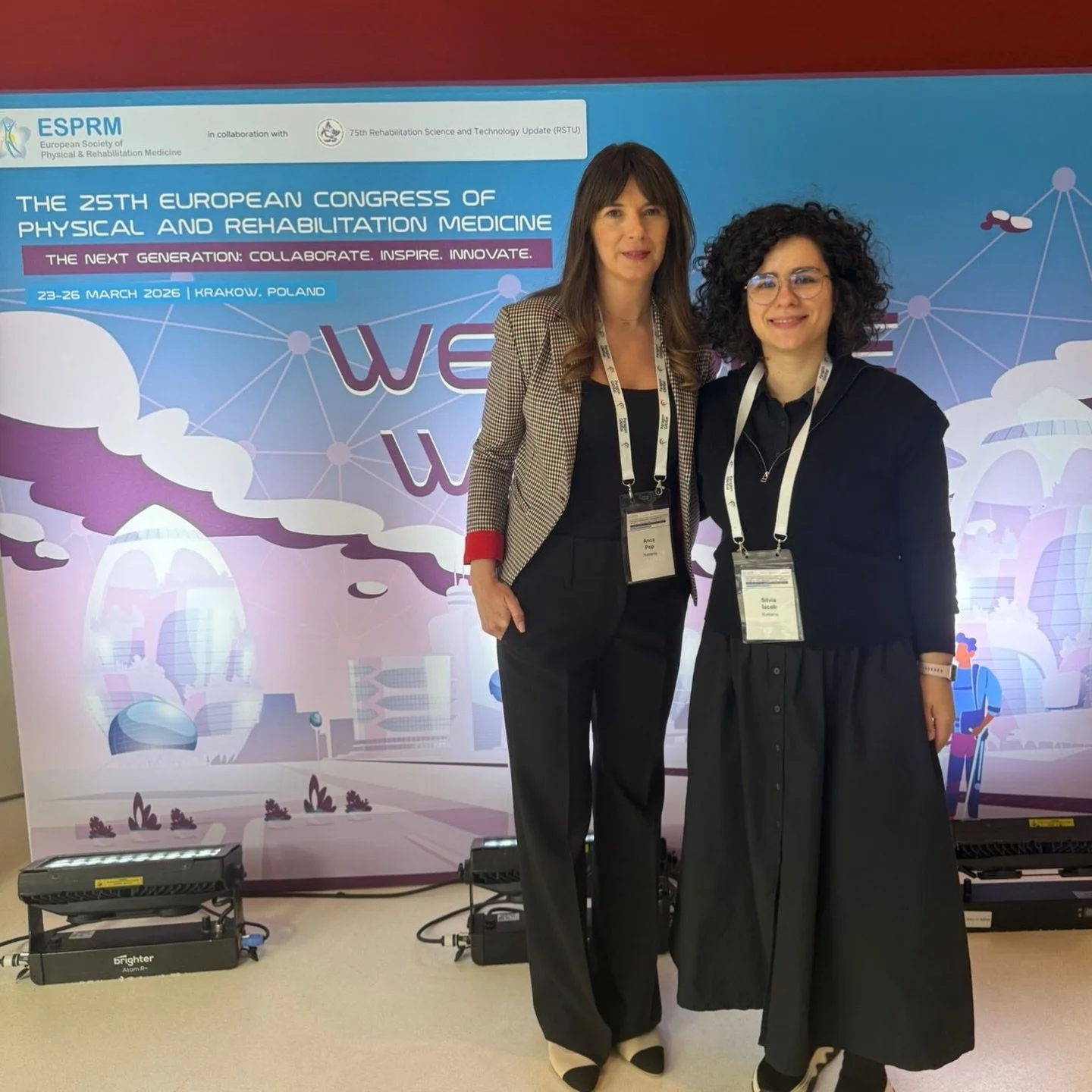 Two women standing in front of a conference backdrop for the European Congress of Physical and Rehabilitation Medicine, smiling at the camera. They are wearing conference badges and lanyards, with cityscape icons and conference details on the backdrop.