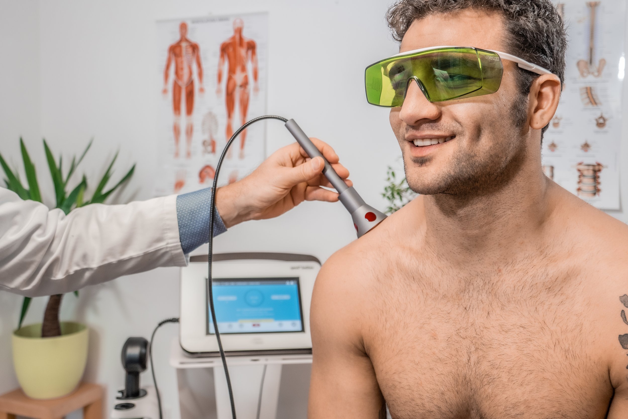A man with curly hair wearing yellow-tinted protective glasses is having his chest examined with medical equipment by a healthcare professional. Medical posters and a computer are visible in the background.