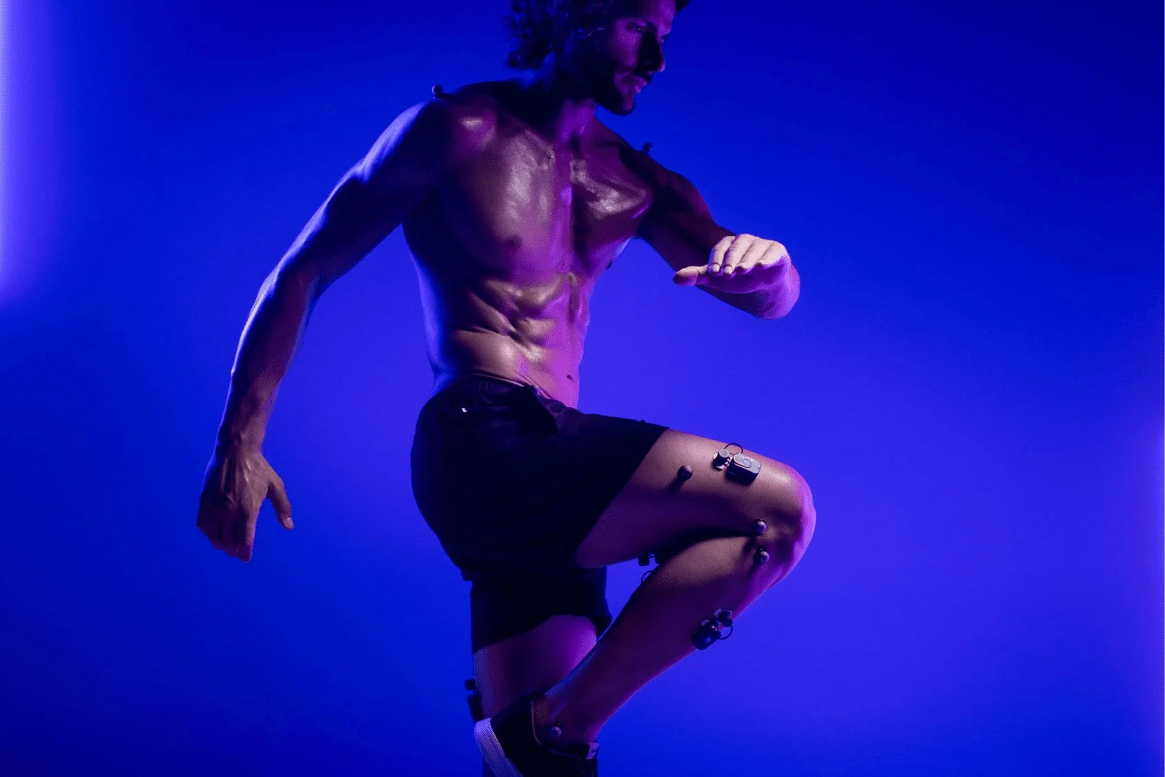 A shirtless man with curly hair and a beard, wearing black shorts, is in a crouched position against a purple background. He has electronic sensors attached to his leg and body, possibly for motion analysis or medical monitoring.
