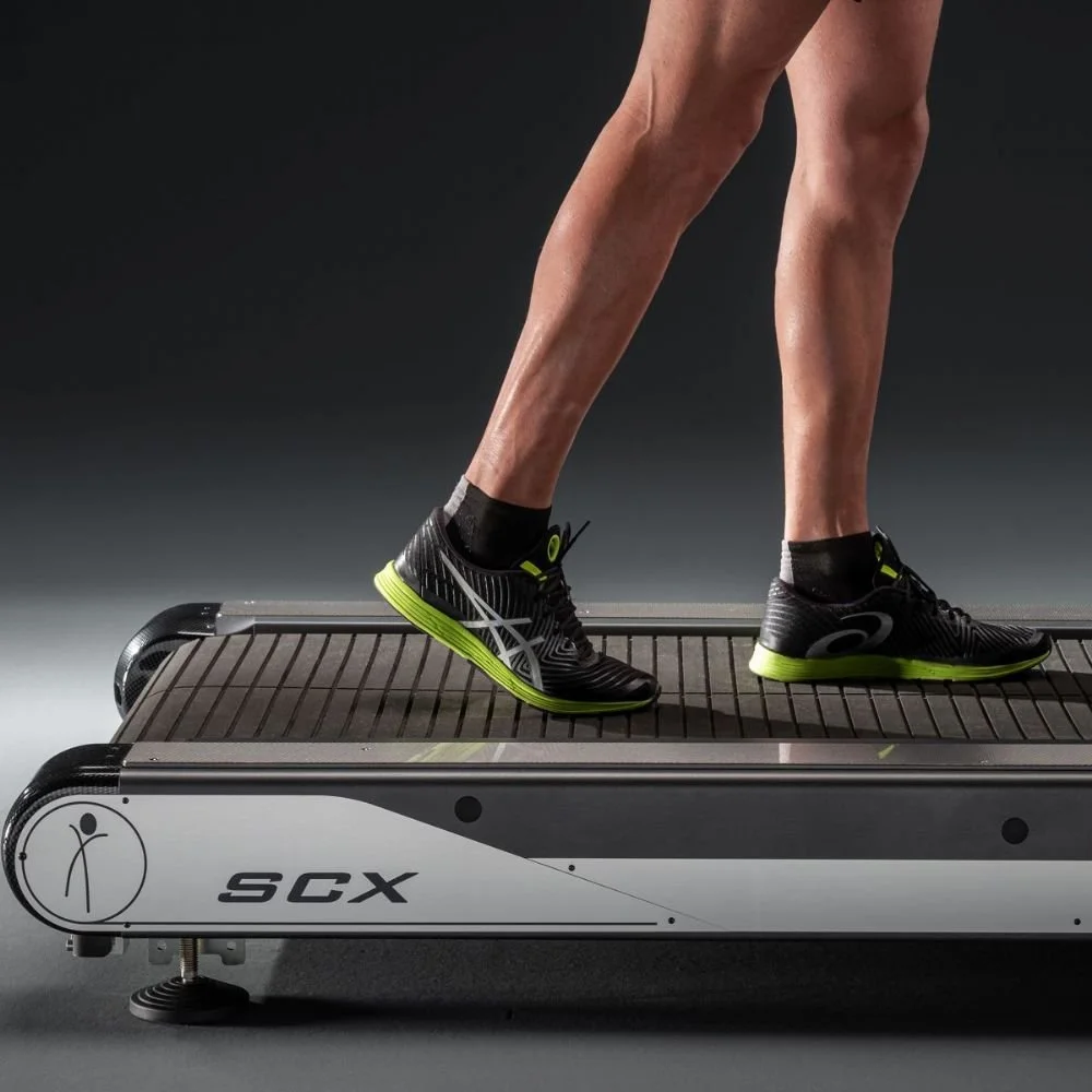 A person jogging on a treadmill with black running shoes with green soles, wearing black socks, in a dark studio setting.