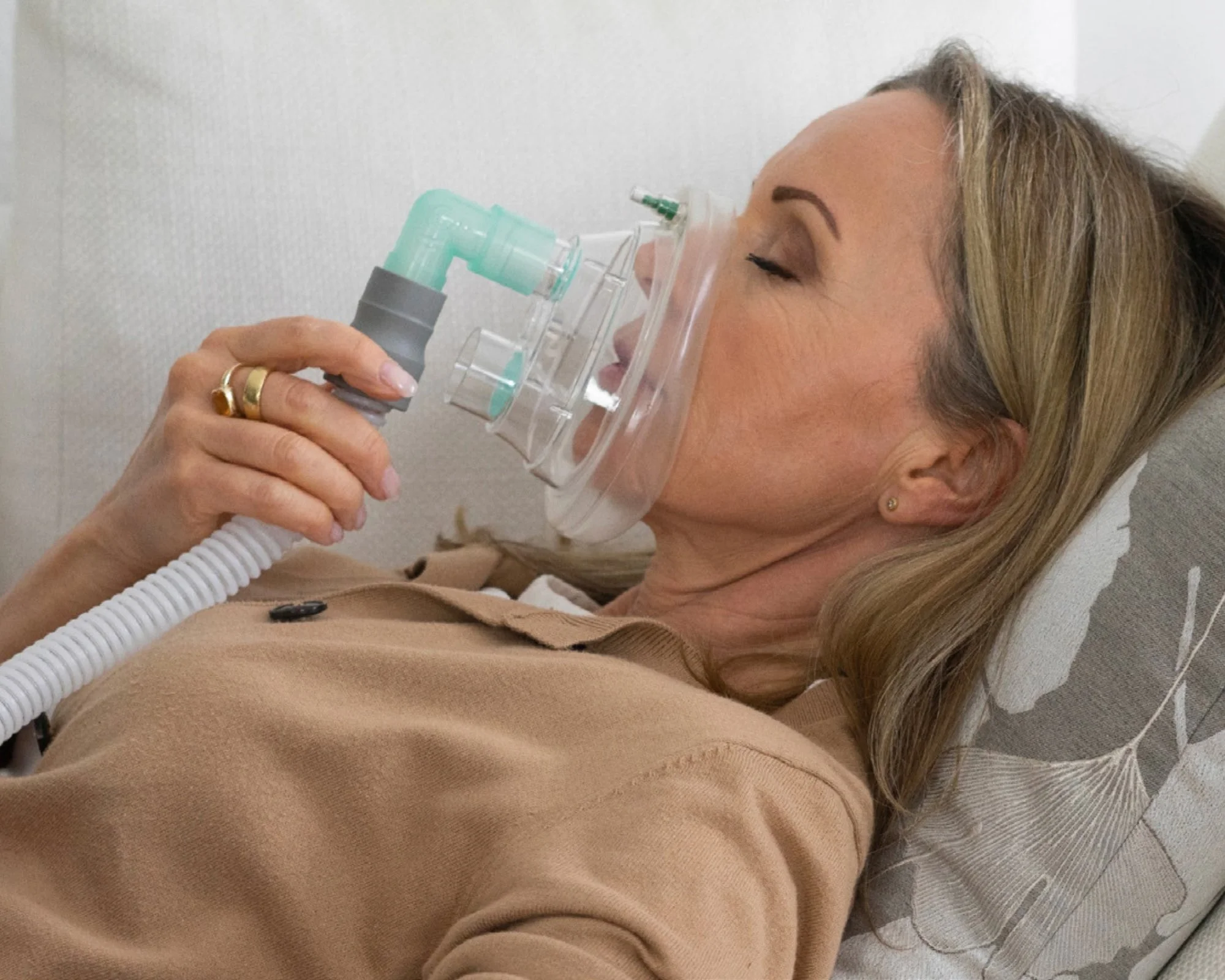 A woman with blonde hair and earrings lies on a medical bed, using a nebulizer with a mask on her face.