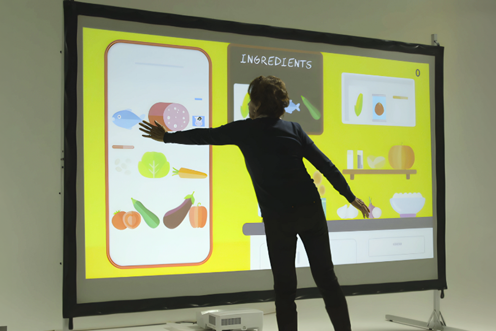 Person interacting with a large digital screen displaying a recipe app with ingredients including fish, cheese, carrots, green leaves, zucchini, eggplant, and pumpkins.