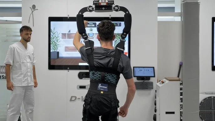 A man is wearing a robotic exoskeleton on his arms while standing in front of a large screen in a medical or rehabilitation setting. A nurse or medical professional in white scrubs observes nearby.