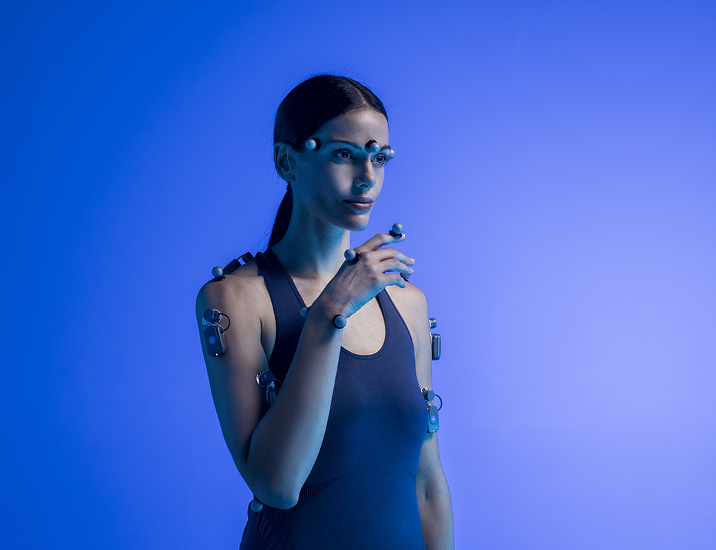 A woman with long dark hair wearing a black athletic top, covered in sensors and electrodes, stands against a blue background in a technology and innovation concept.