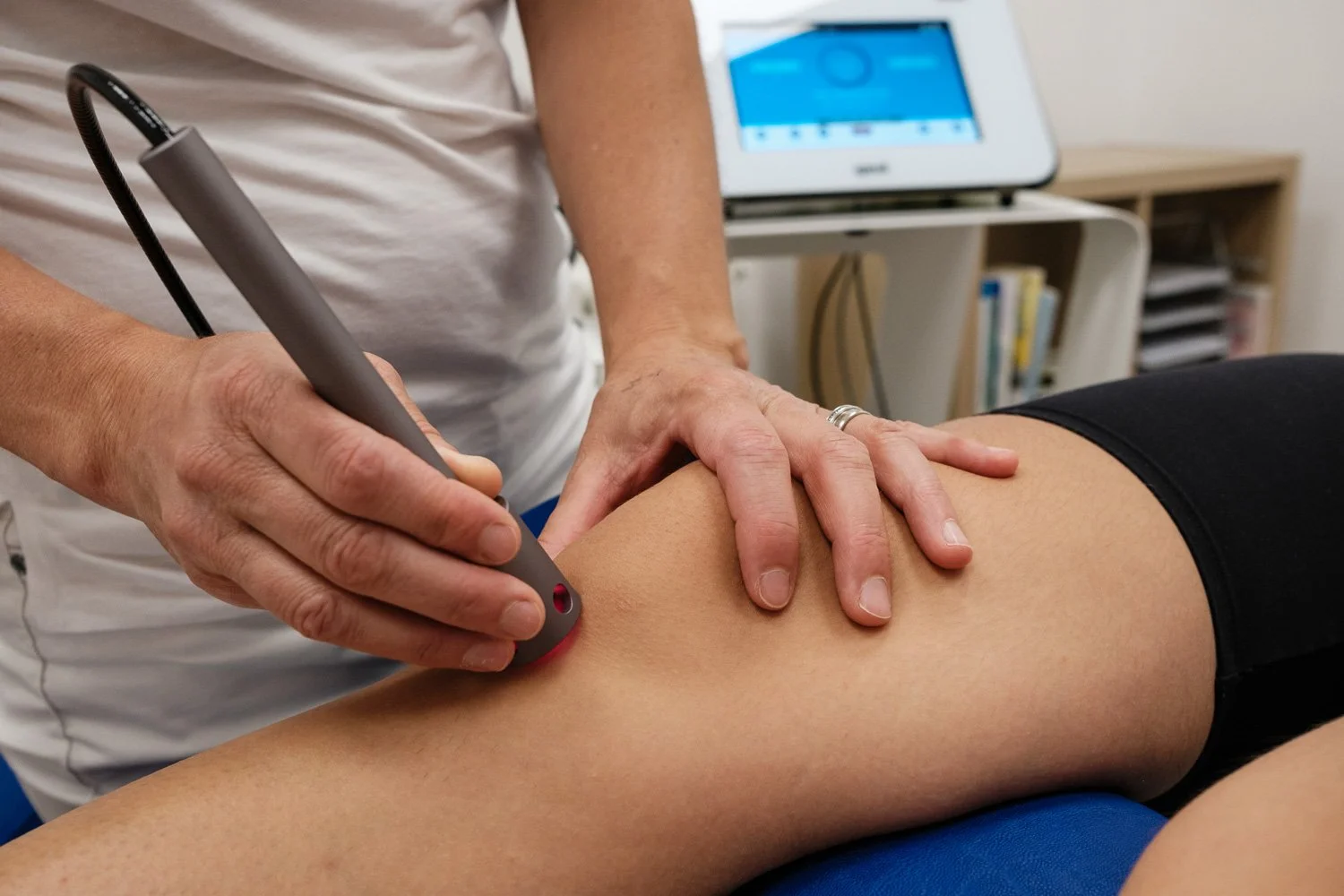A healthcare professional is performing a medical procedure on a patient's knee using a specialized device, with a monitor in the background.