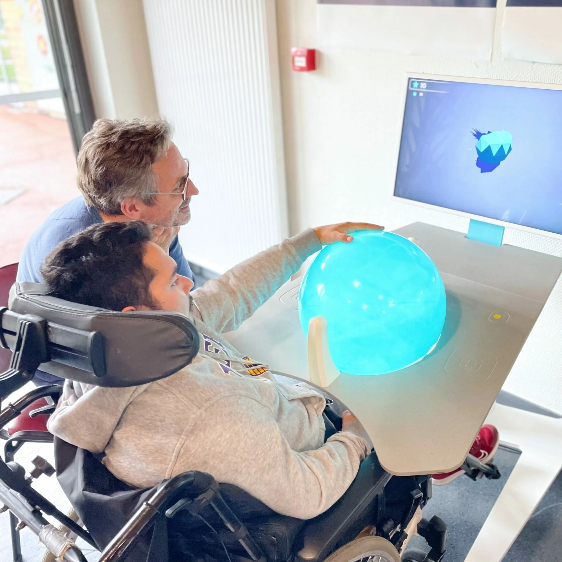A person in a wheelchair, wearing a hoodie, is playing a video game on a computer with a large monitor. There are two children beside him, one with short dark hair and the other with lighter hair, watching and engaging with him. The person is holding a glowing blue ball, and the video game on the monitor features a low-poly dinosaur or creature.
