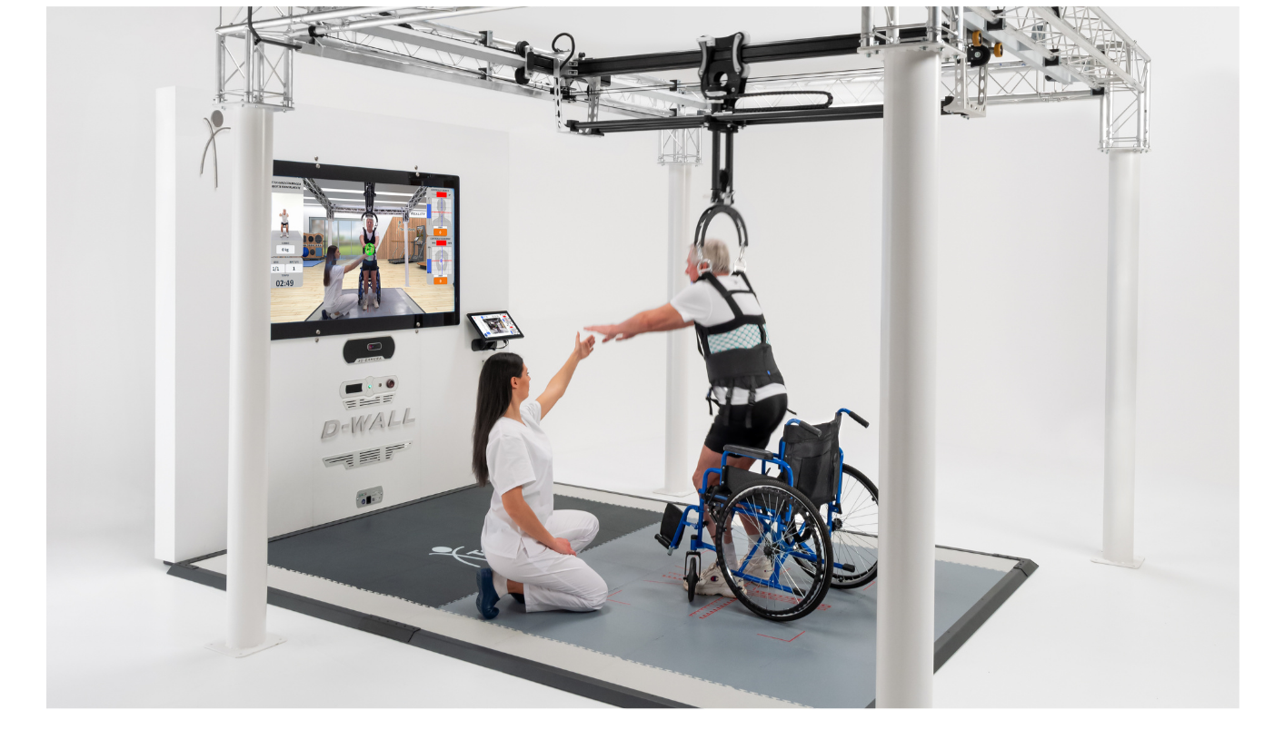 A healthcare professional assists a person in a wheelchair to perform a rehabilitation exercise using a robotic device. The person in the wheelchair is reaching out to a trainer who is kneeling on a mat, with both of them pointing towards a large monitor displaying the exercise session. The setting is a modern, white medical or rehabilitation facility with advanced equipment.