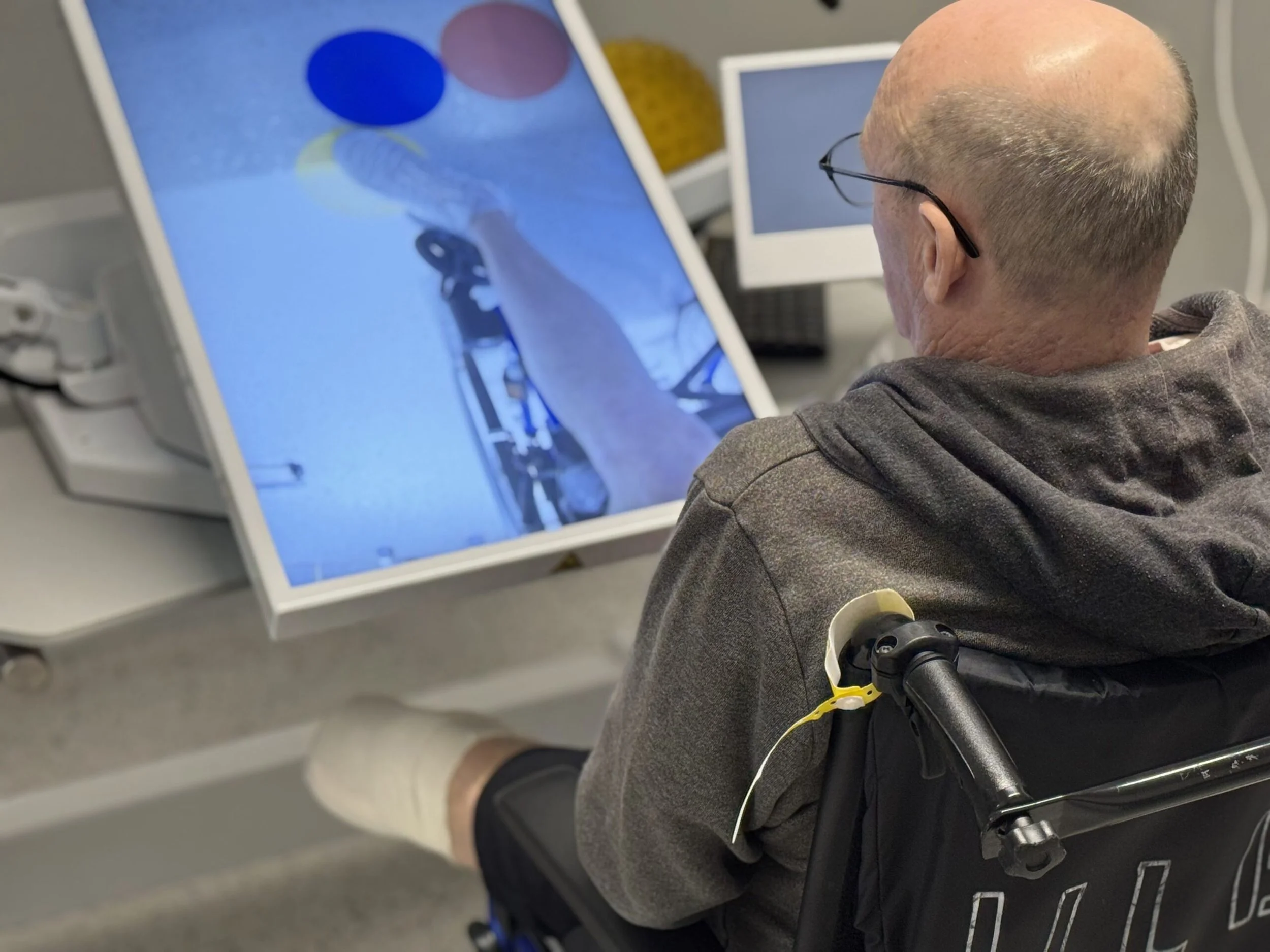 A man with a shaved head and glasses sitting in a wheelchair, using a touchscreen computer on a desk, with a keyboard and wires nearby, in an indoor setting.