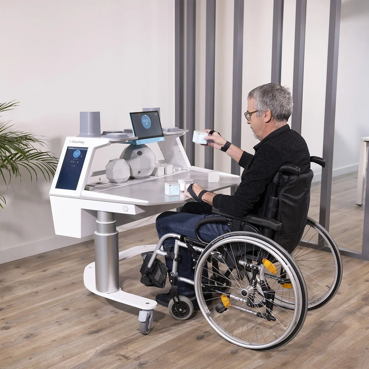 A man in a wheelchair sitting at a desk with a computer and some objects, holding a small box, in a modern, minimalist office or laboratory setting.