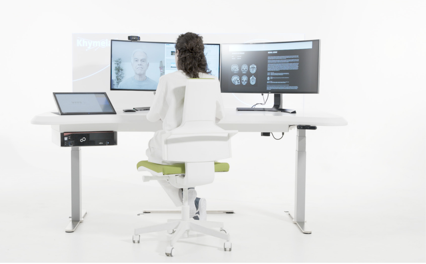 Medical professional in a white coat sitting at a white desk with three monitors, engaging in a telemedicine consultation with a patient, in a bright, minimalistic room.
