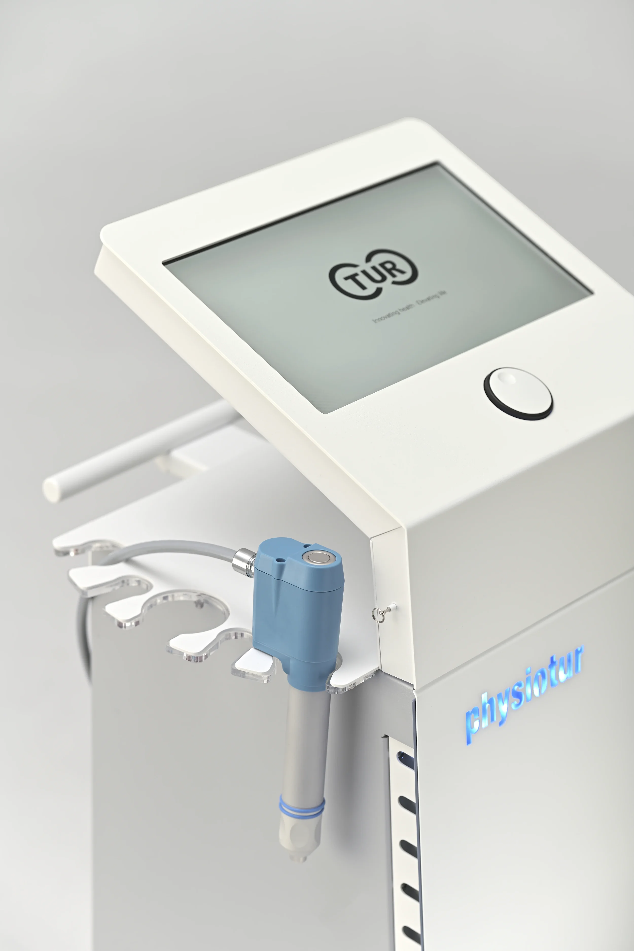 A medical device with a touchscreen display that reads "TUR" and has a stylus attached, positioned on a white stand with a row of circular holes and blue text that says "physiotur".