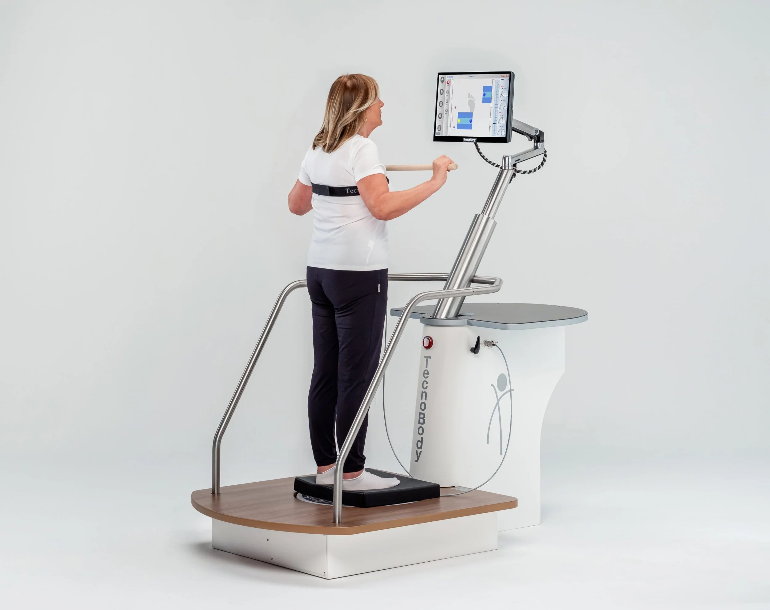 A woman is standing on a digital balance platform in a medical or testing device, holding a wooden handle and looking at a screen showing body or health data.